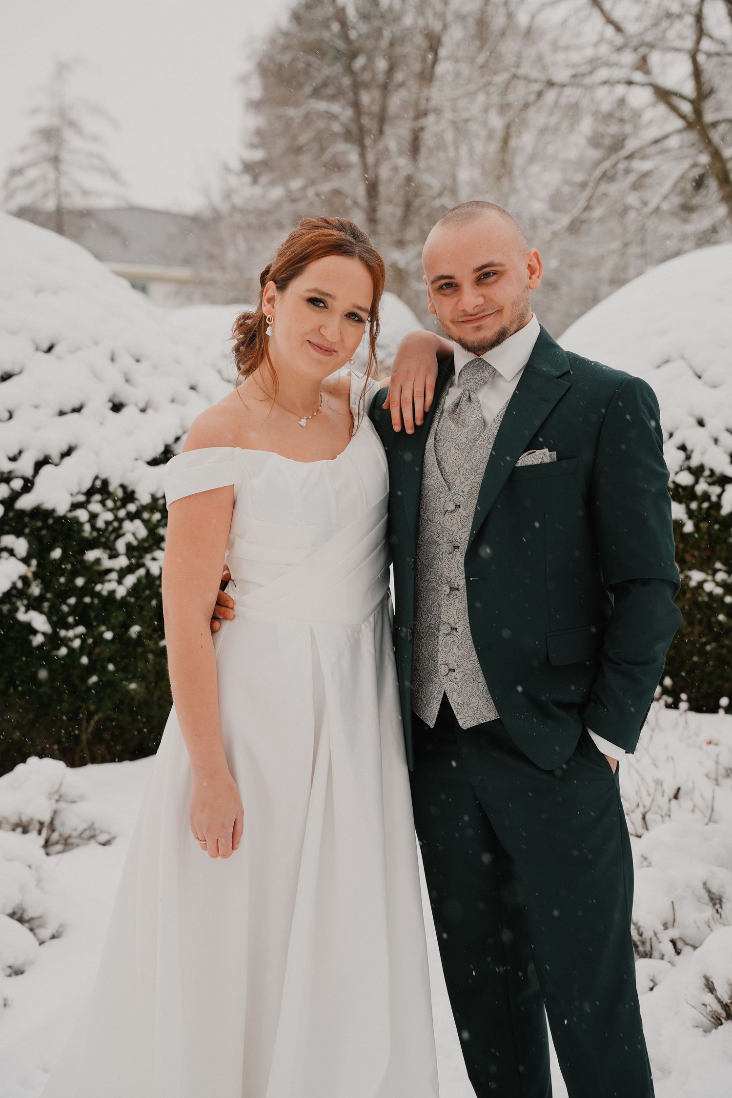A bride and groom pose together in a snowy winter garden, she in a white gown, he in a dark green suit.