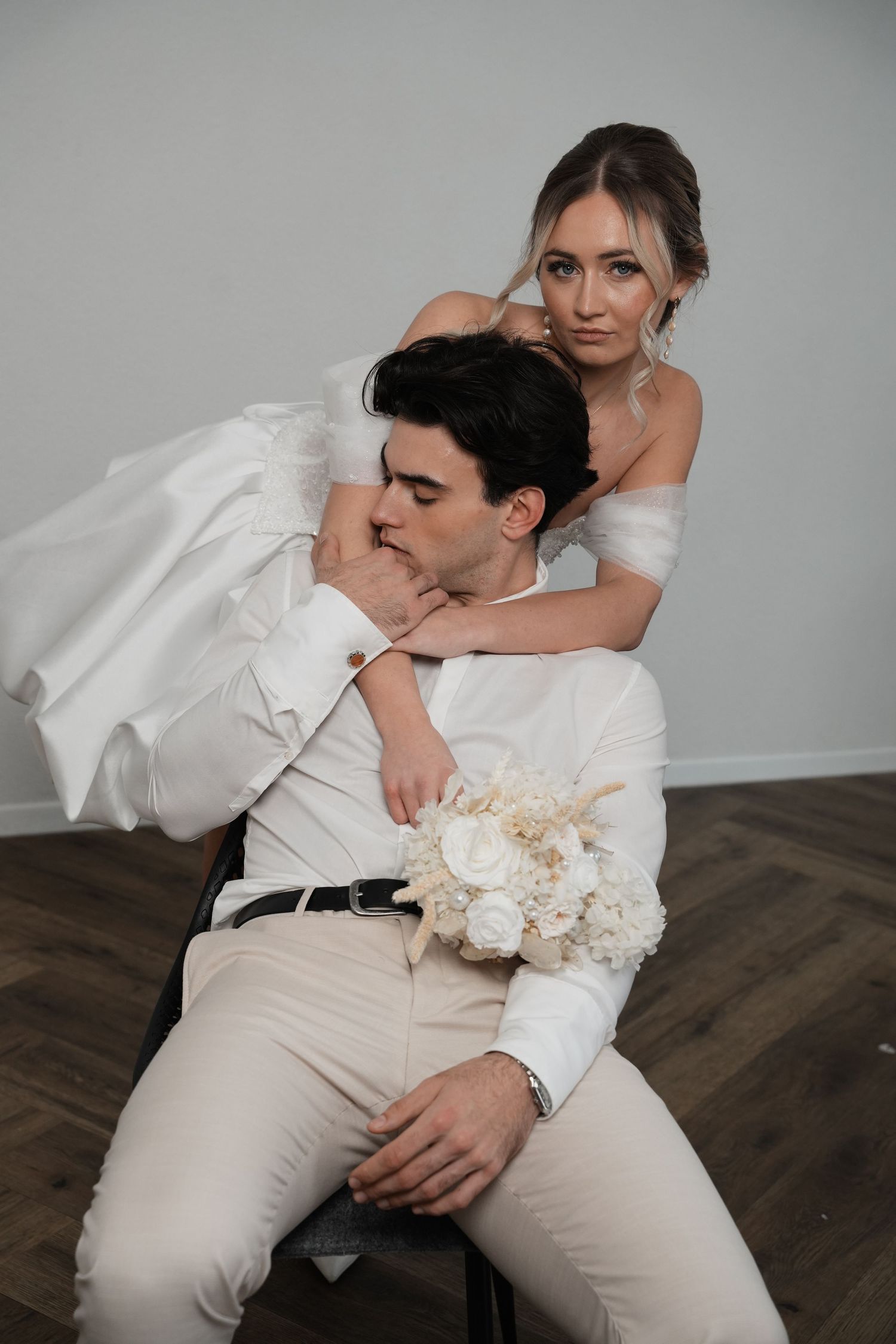 Bride in white off-shoulder gown embraces groom in white suit holding a bouquet during an elegant wedding portrait.