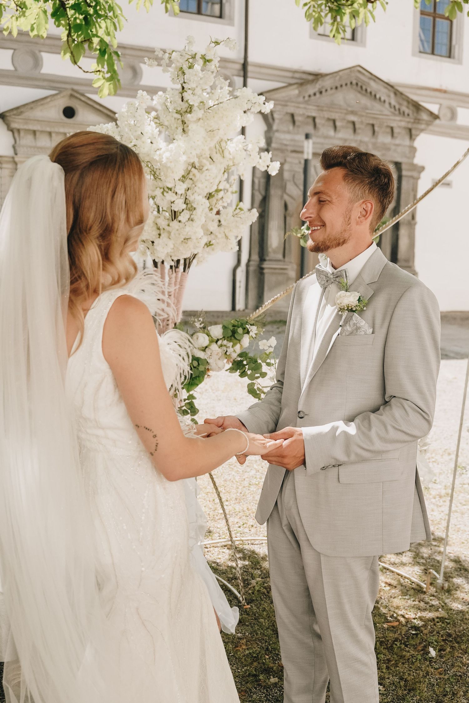 Bride and groom exchanging vows outdoors under white floral arch at elegant wedding ceremony.