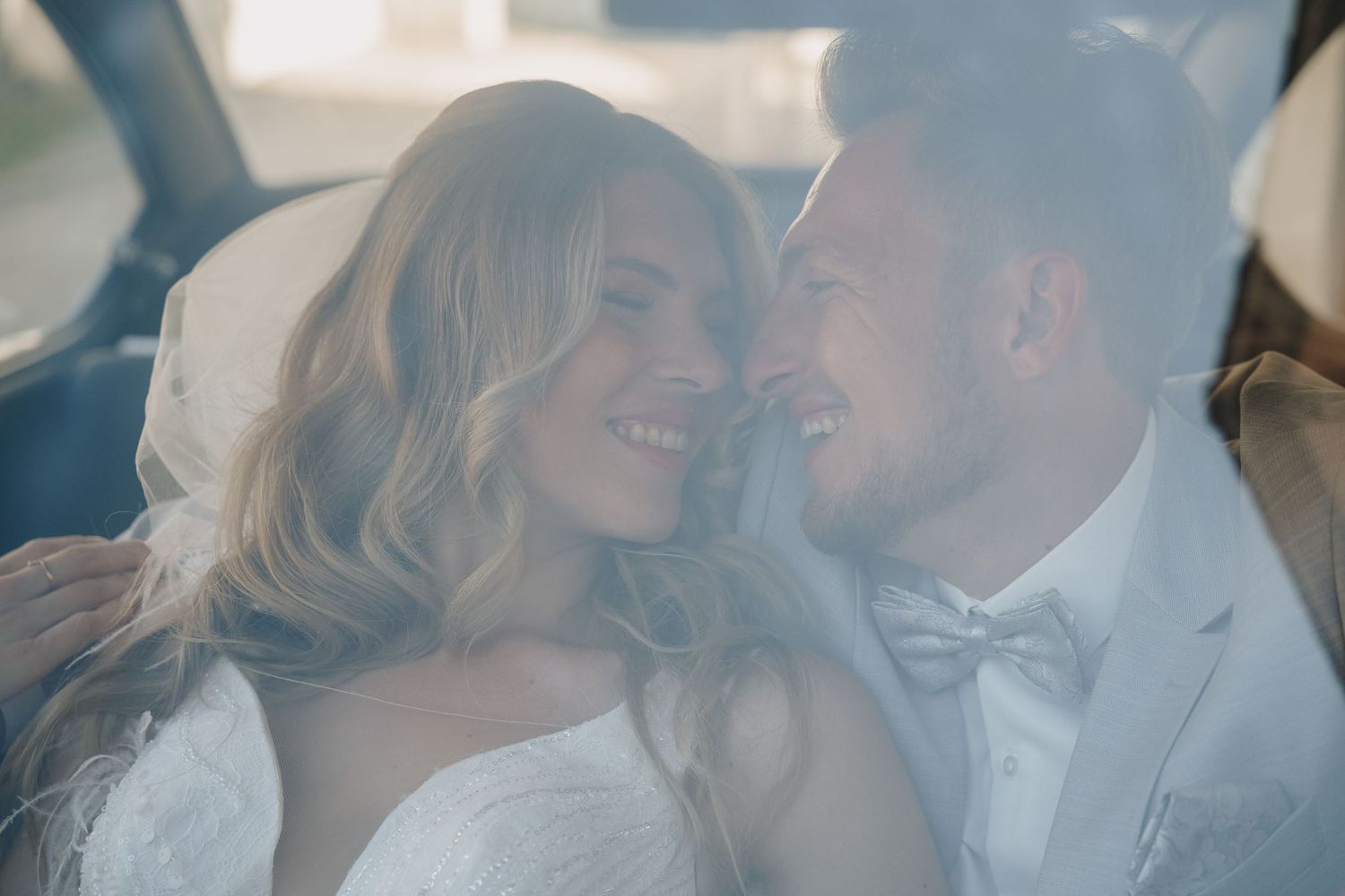 Happy newlyweds share an intimate moment in a car, bride in white dress and groom in white suit with bow tie.