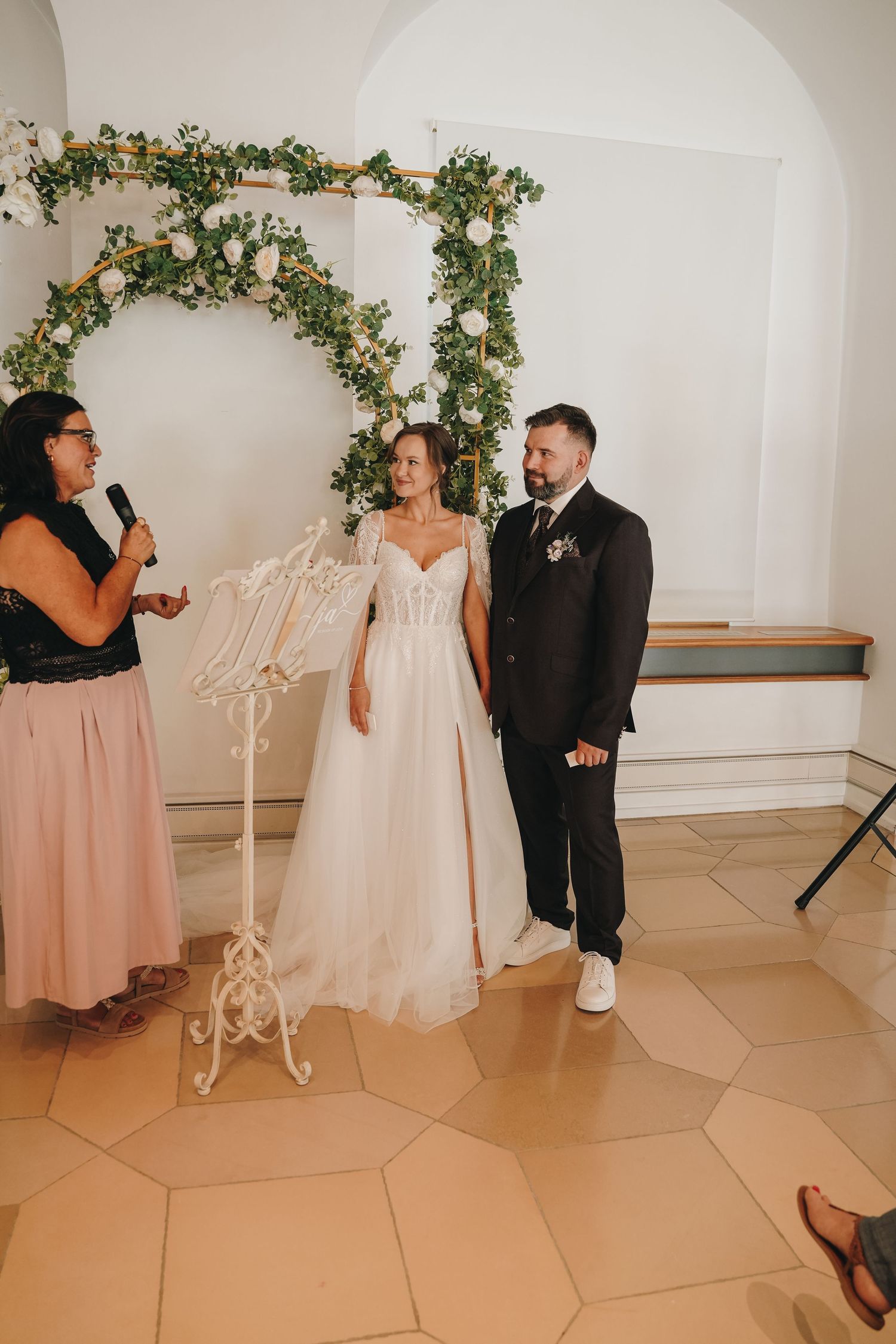 Bride and groom stand before floral arch during indoor wedding ceremony while officiant reads vows.