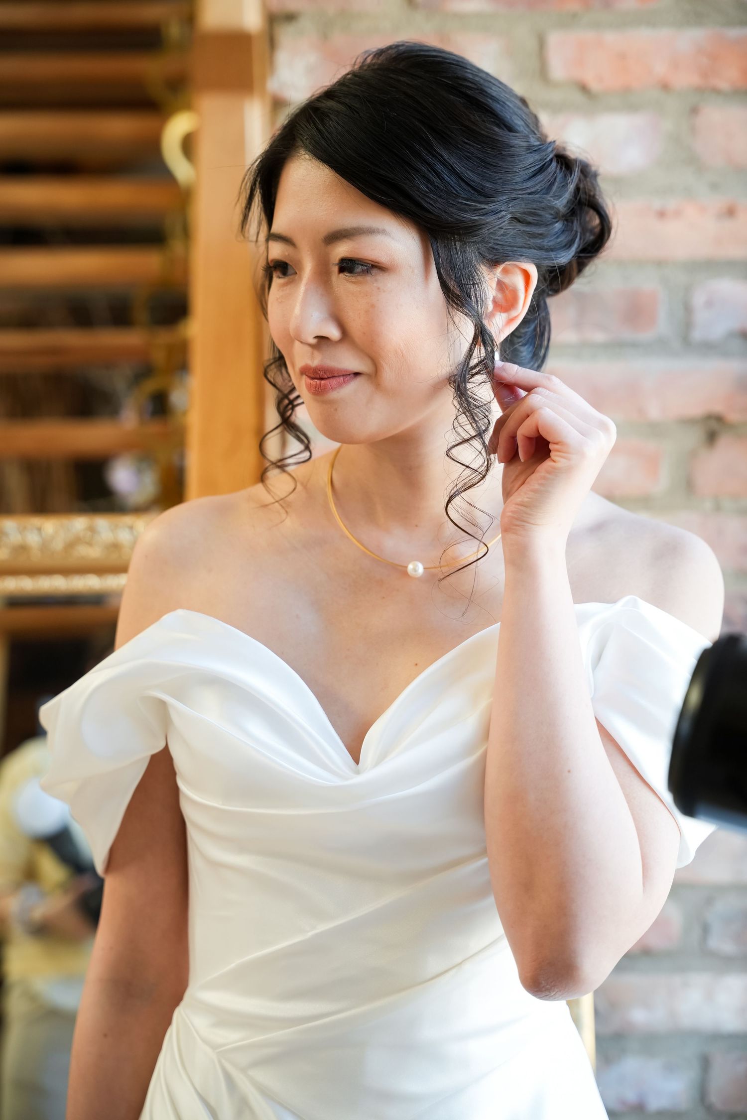 Bride in off-shoulder white gown adjusting pearl earring, wearing gold necklace, with elegant updo hairstyle.
