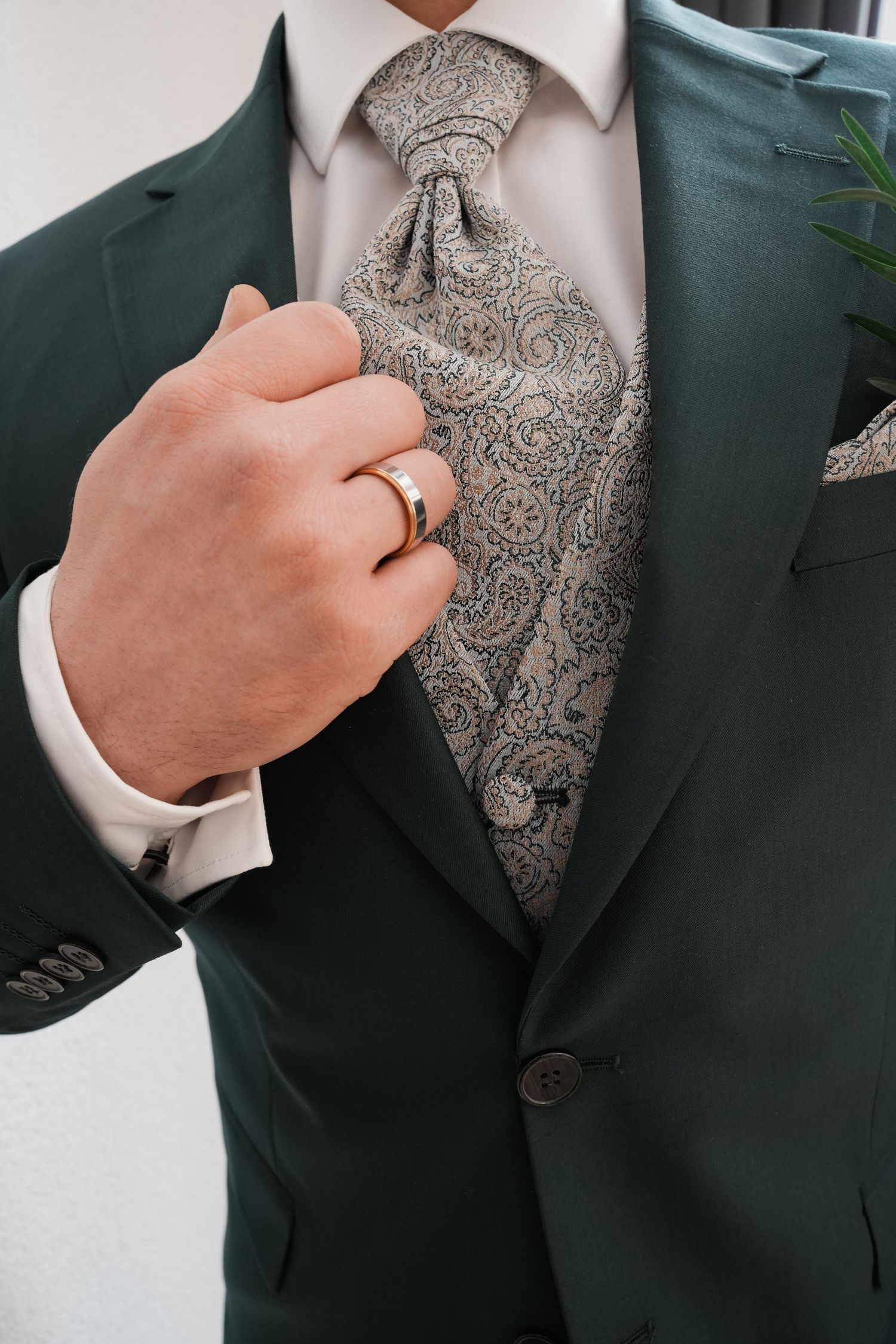 Man in green suit adjusting paisley tie and vest, wearing gold ring, with boutonniere, formal wedding attire.