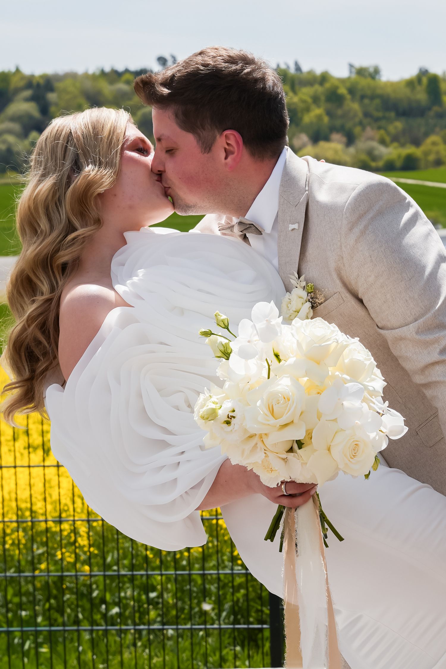 Bride and groom share a romantic kiss, bride holding white rose bouquet, outdoors with green fields backdrop.