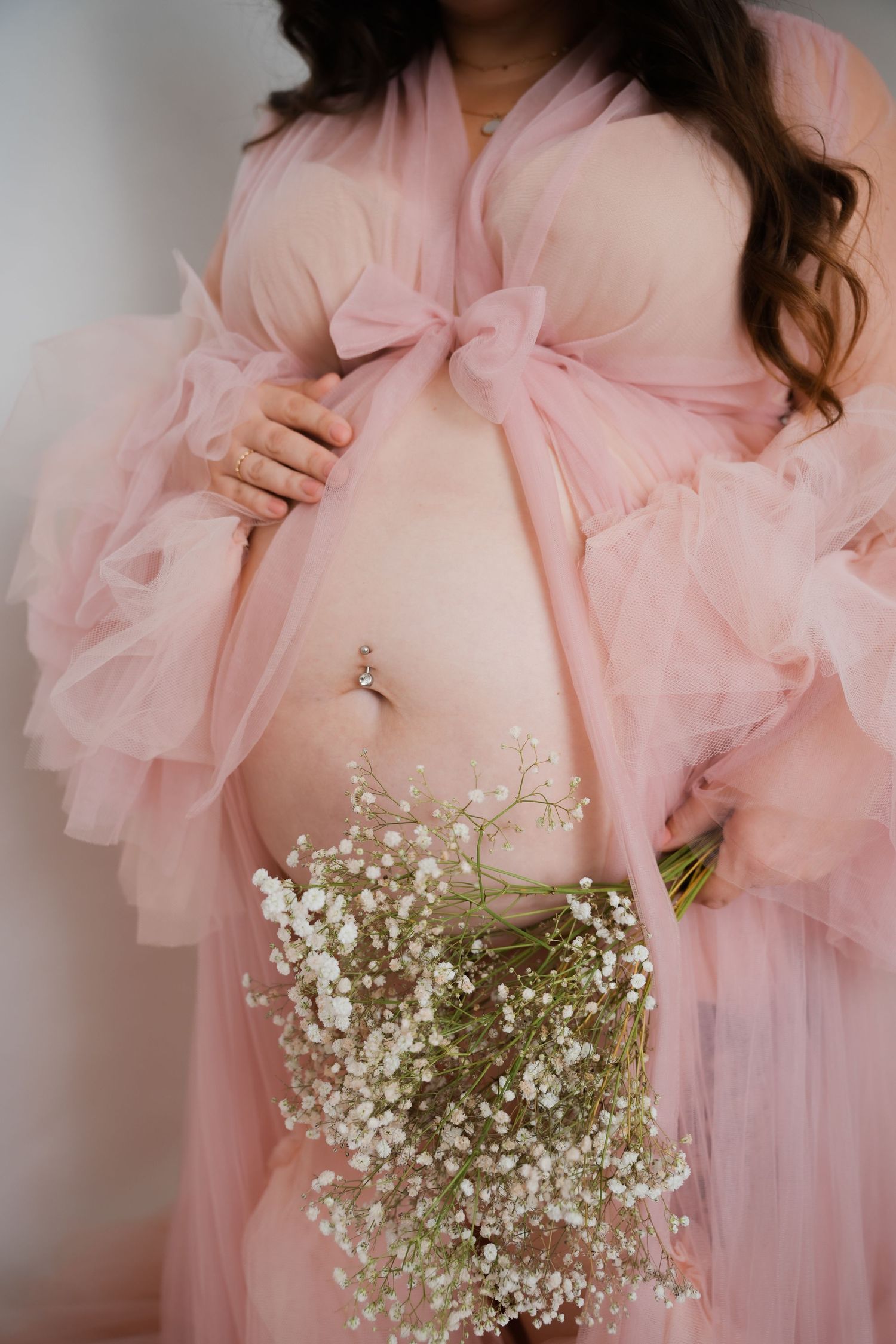 Pregnant woman in pink tulle robe holding baby's breath flowers, showcasing baby bump with belly piercing.