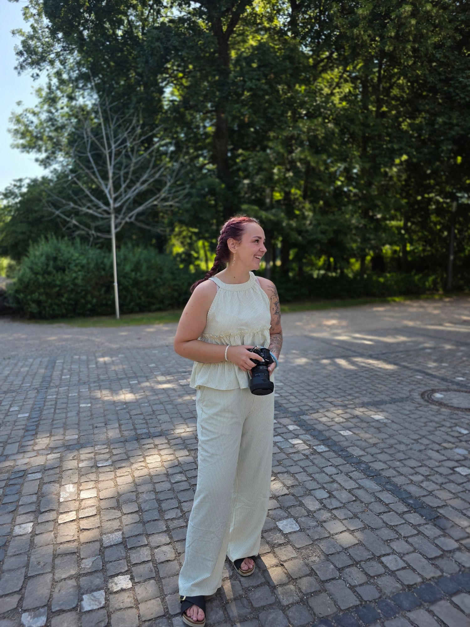 Woman in cream outfit holding a camera stands on cobblestone path surrounded by lush green trees.