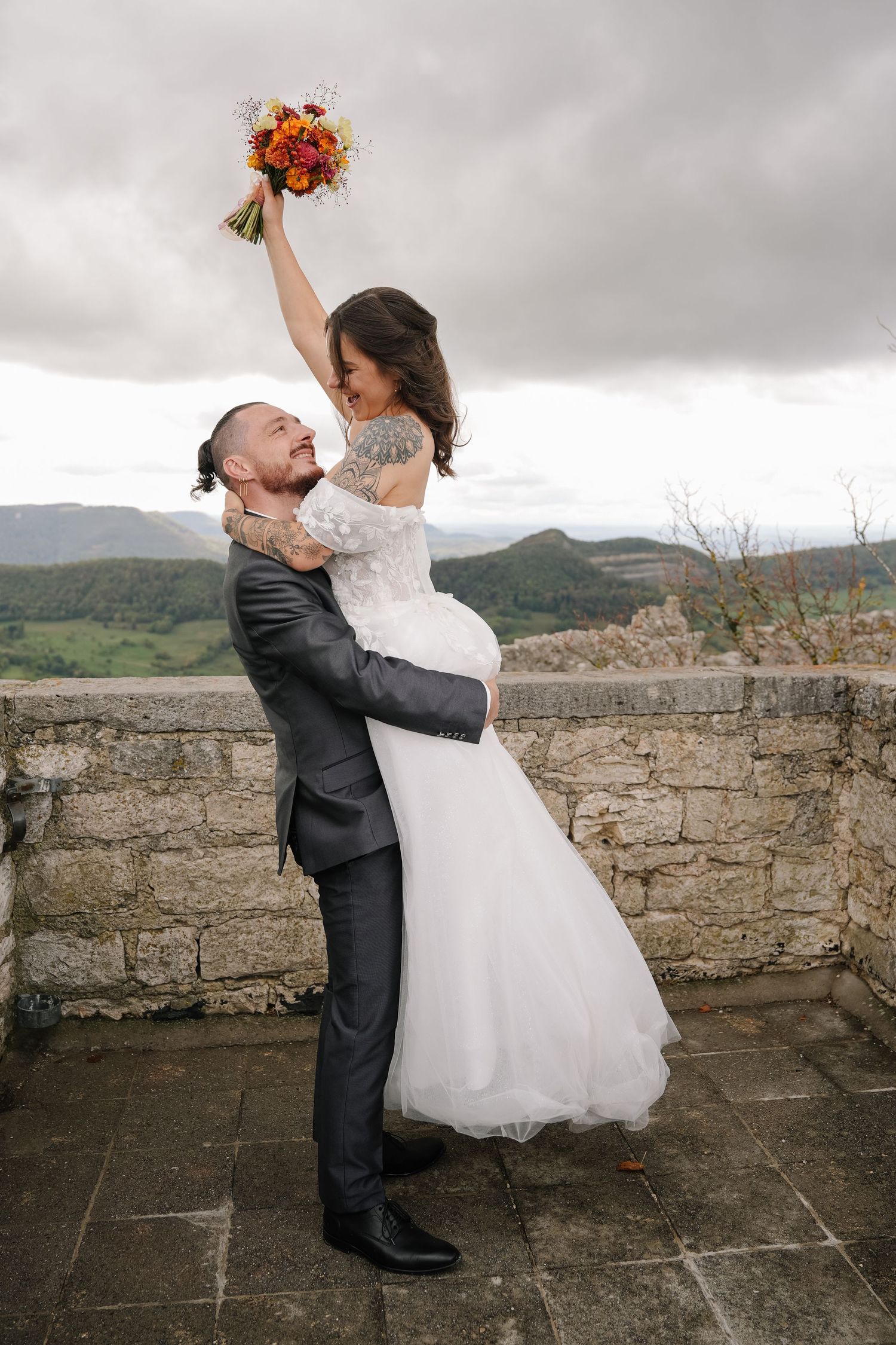 Groom lifts smiling bride in white wedding dress holding an orange bouquet on a stone terrace overlooking green hills under a cloudy sky.