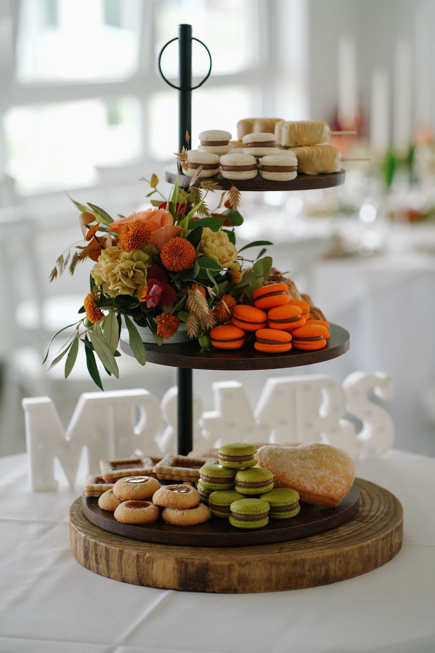 Three-tiered wooden dessert stand with orange and green macarons, cookies, and an autumn floral arrangement on a wedding reception table.
