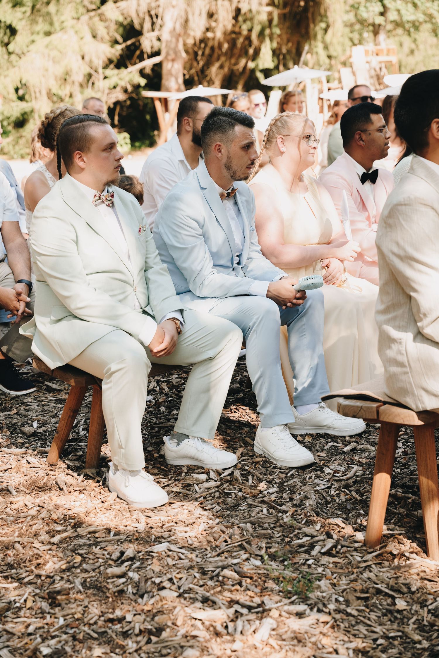 Guests in light suits and white sneakers sit outdoors at a woodland wedding ceremony on a sunny day.