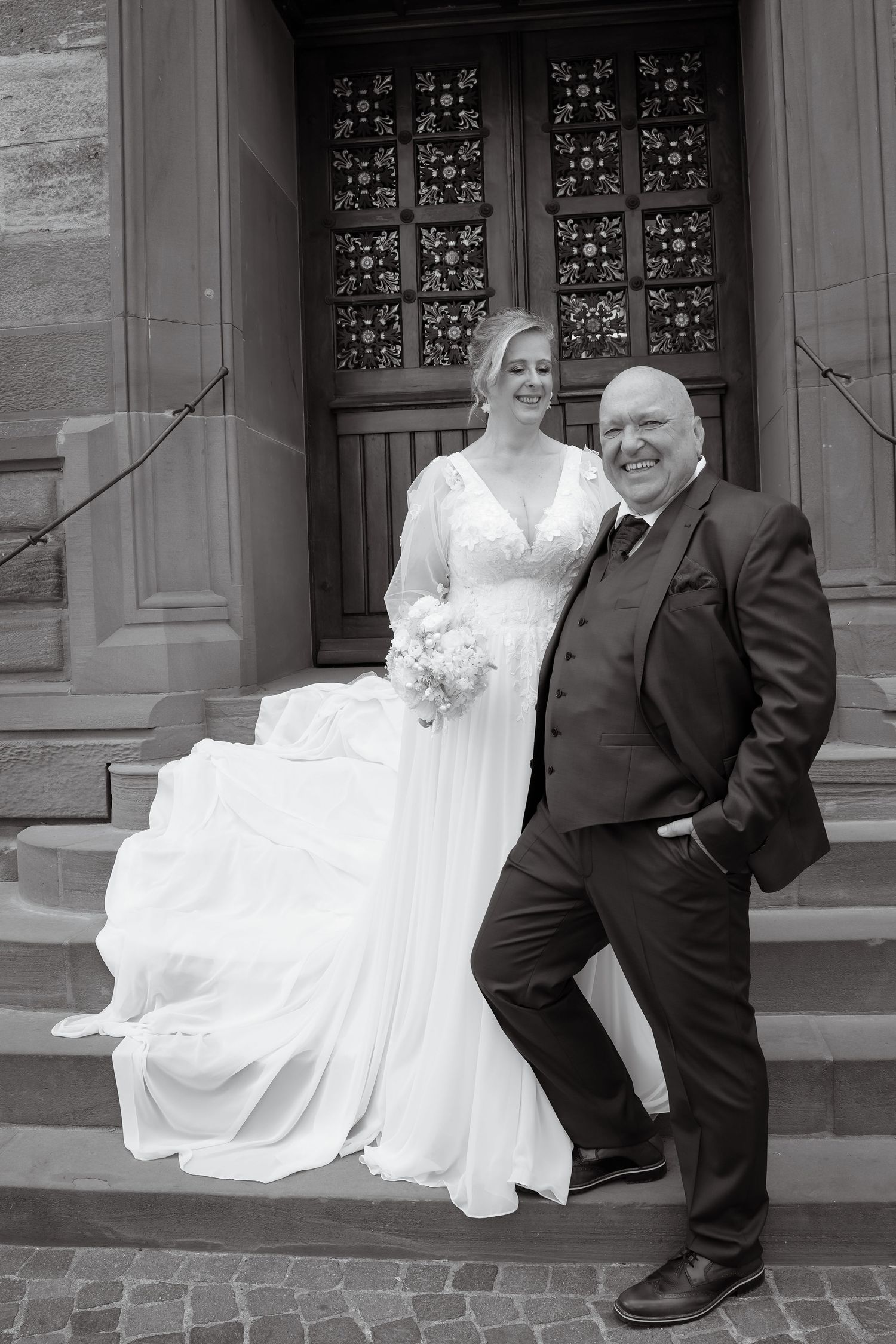 Black and white wedding photo of bride in flowing gown and groom in suit posing on church steps.