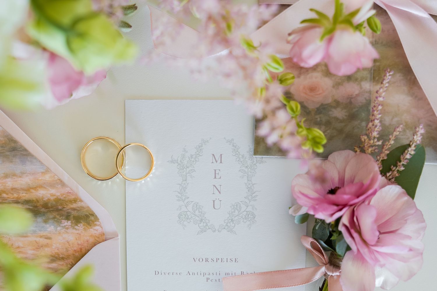 Elegant wedding flat lay with gold rings, pink flowers, and a decorative menu card on a soft pastel background.