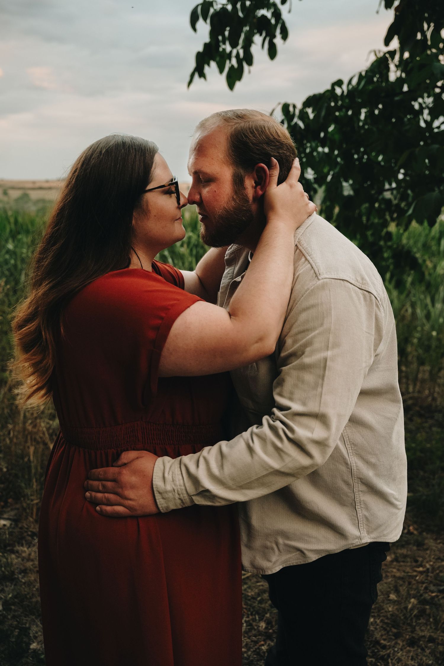 Couple embracing outdoors, woman in red dress touching man's face, romantic engagement photo in natural setting.