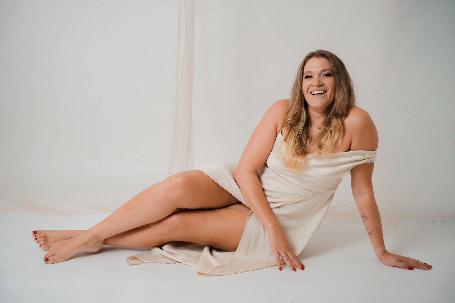 Smiling blonde woman in elegant white off-shoulder dress sitting gracefully on white studio floor.