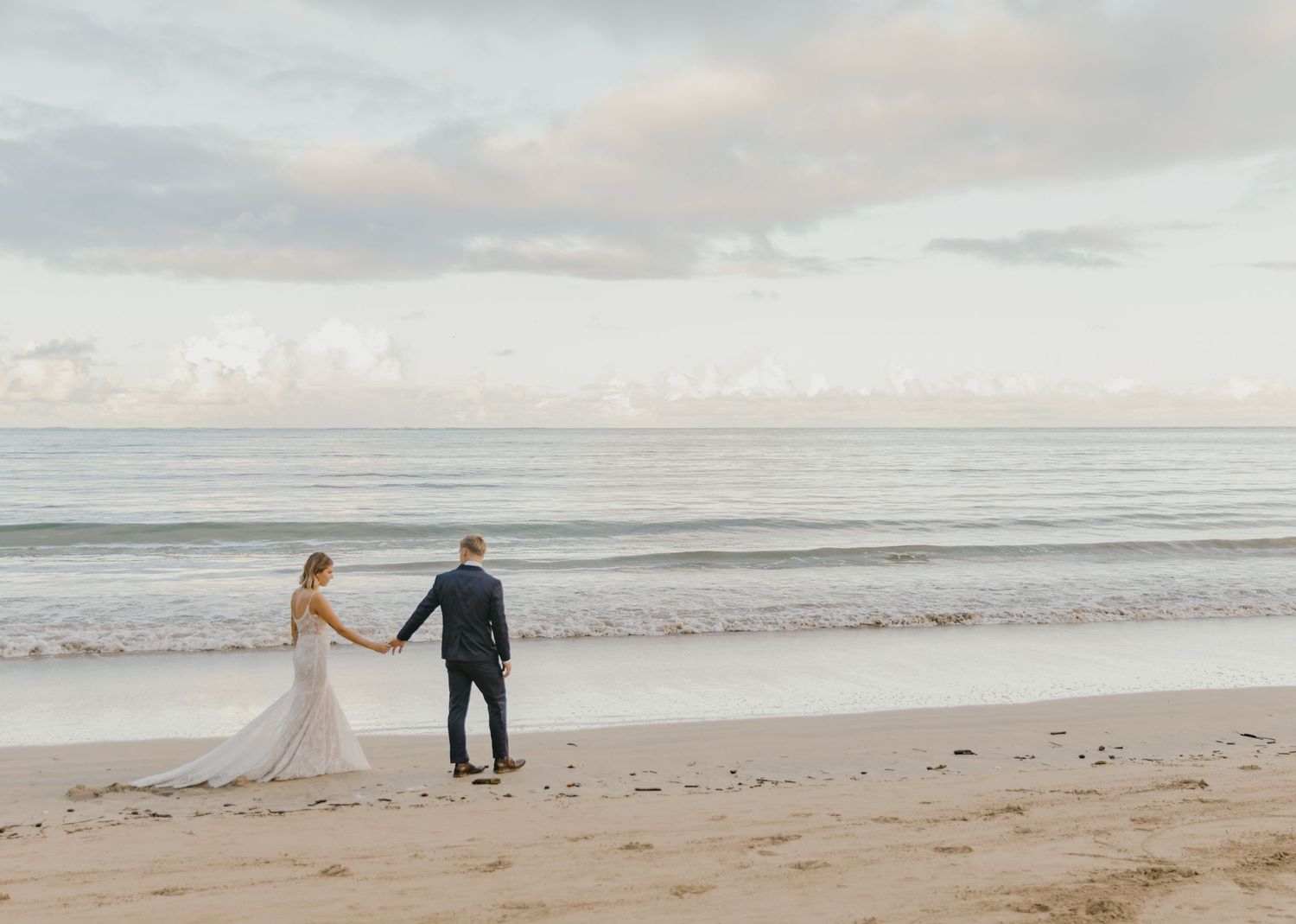 A couple holds hands while walking along a serene beach at sunset with cloudy skies overhead.
