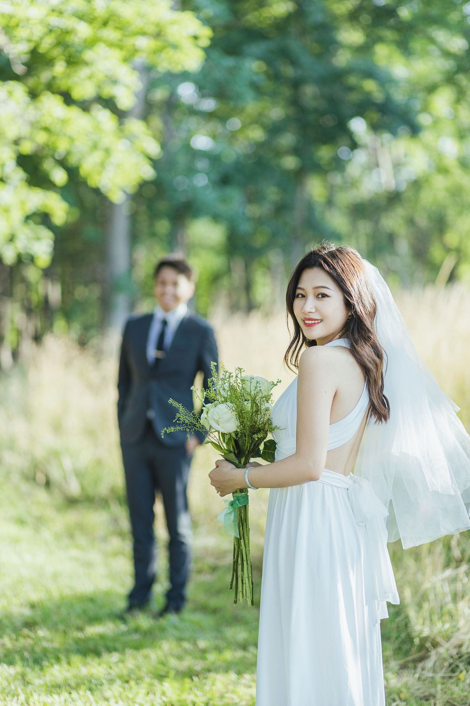A bride in white dress poses with bouquet in sunny garden while groom stands in background.