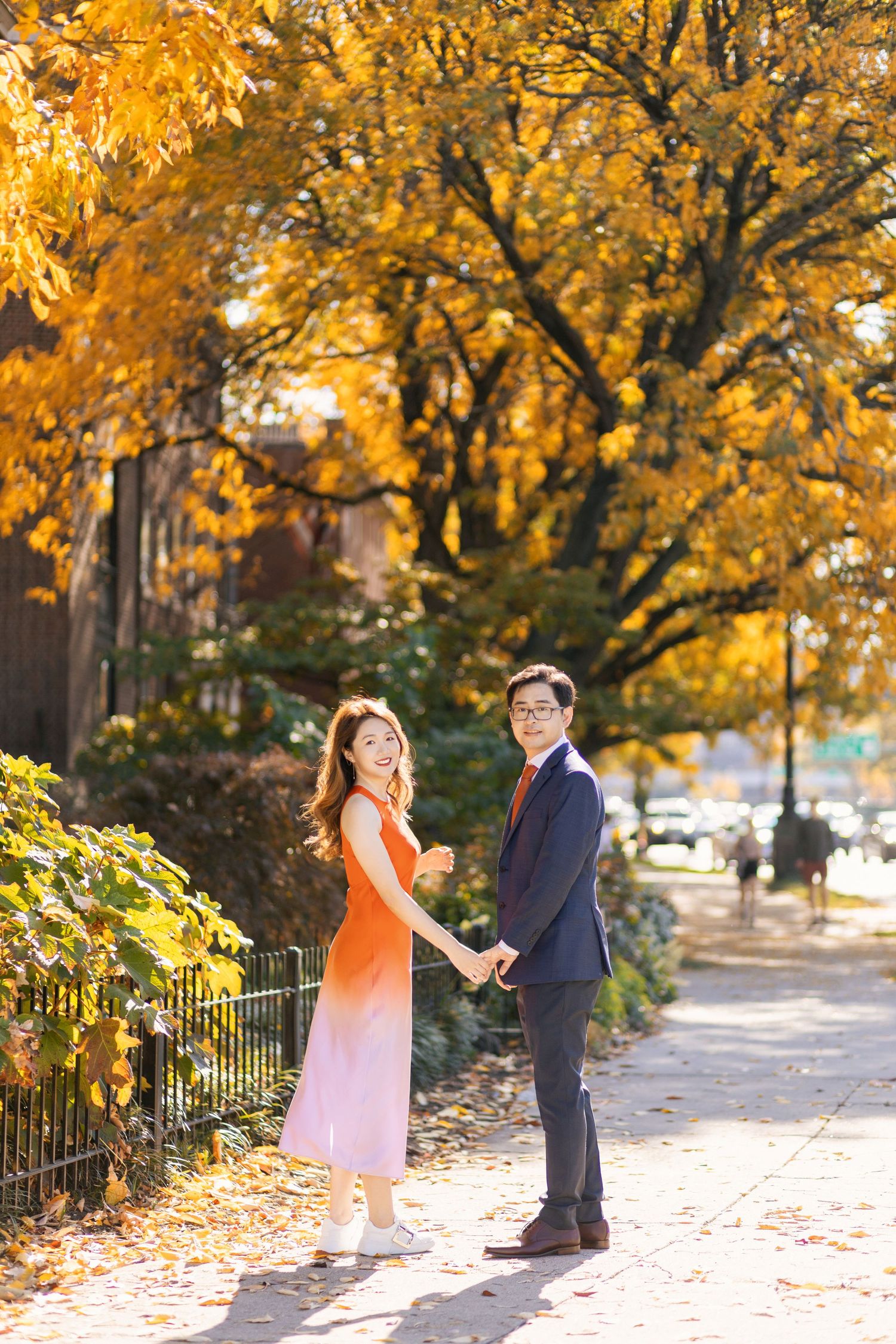 A couple poses on autumn sidewalk surrounded by golden fall foliage.