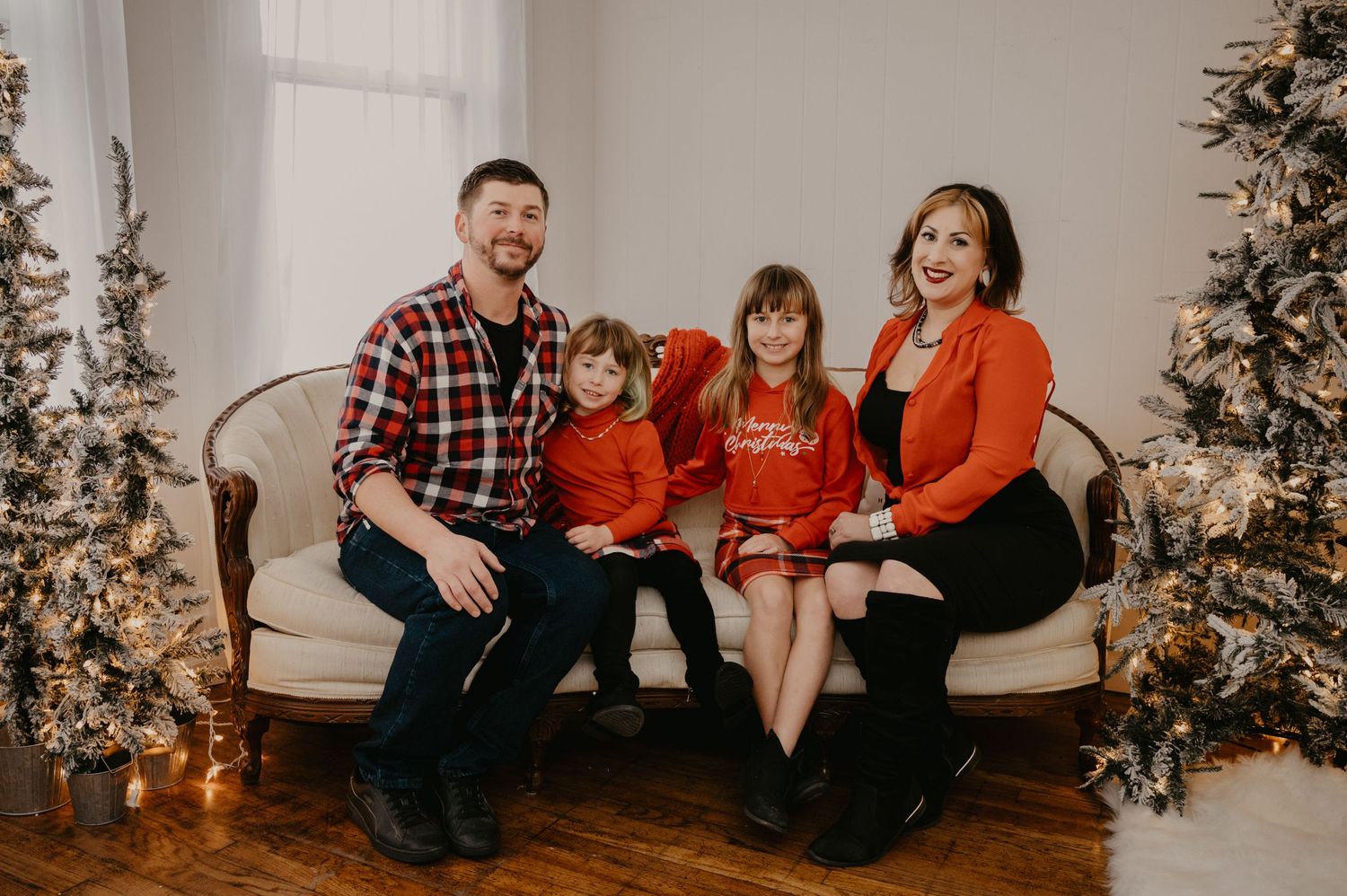 Family of four in red and plaid outfits posing on sofa between decorated Christmas trees.