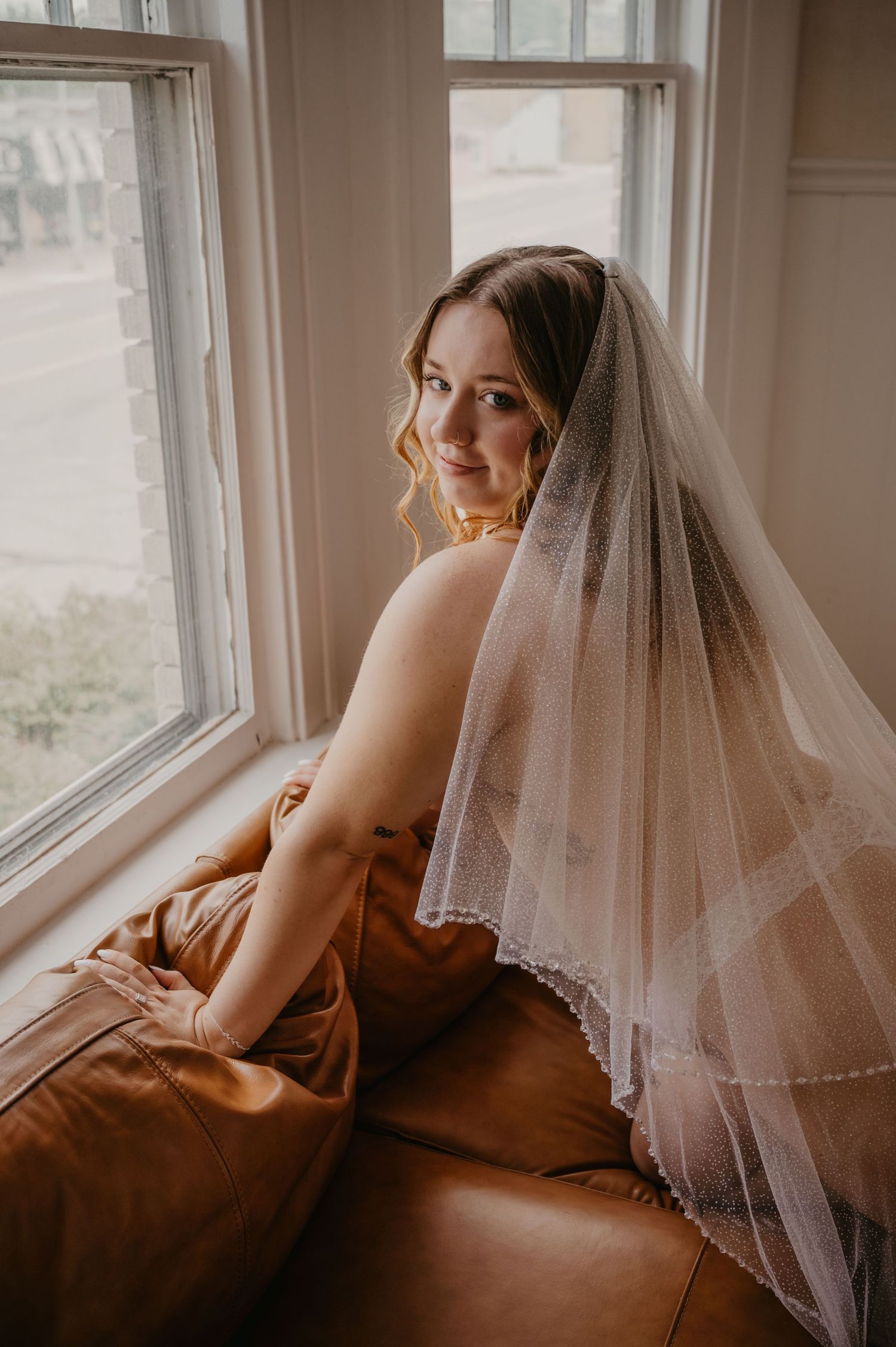 Bride wearing a delicate white veil sits on a leather couch by a sunlit window, smiling over her shoulder.