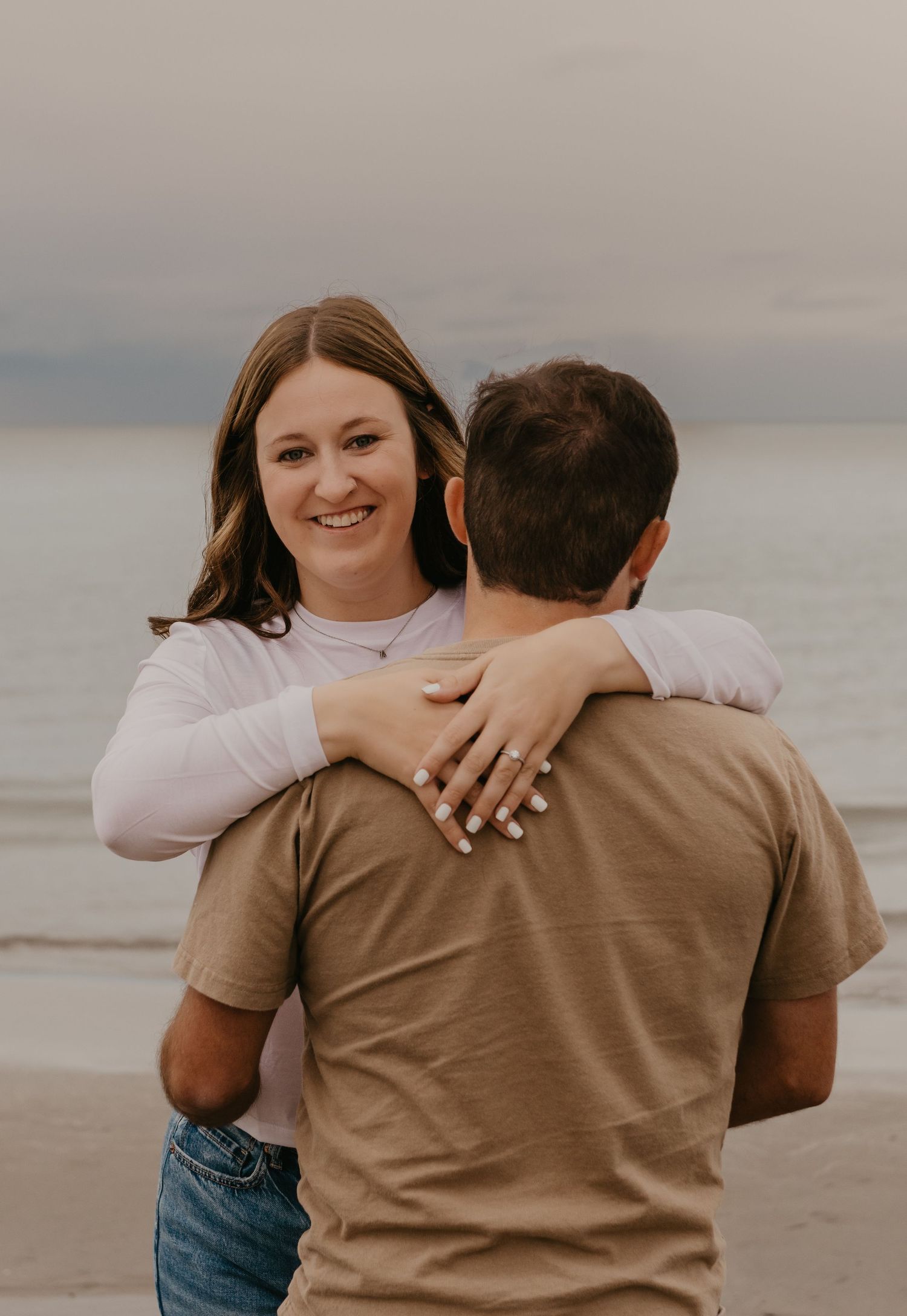Smiling woman hugging her partner on beach at sunset showing engagement ring on her hand.