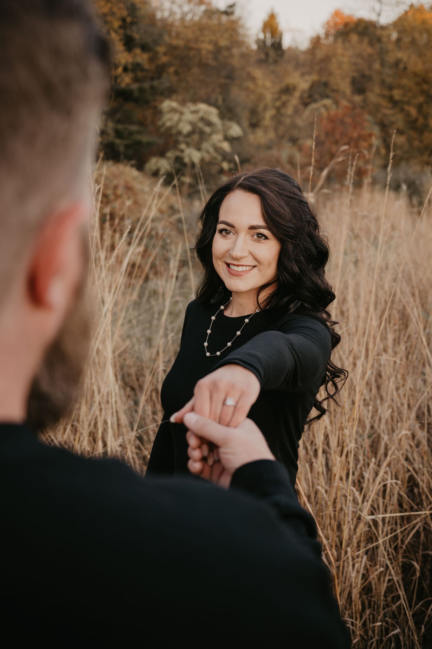A smiling dark-haired woman in a black outfit holds hands with her partner in a golden autumn field.