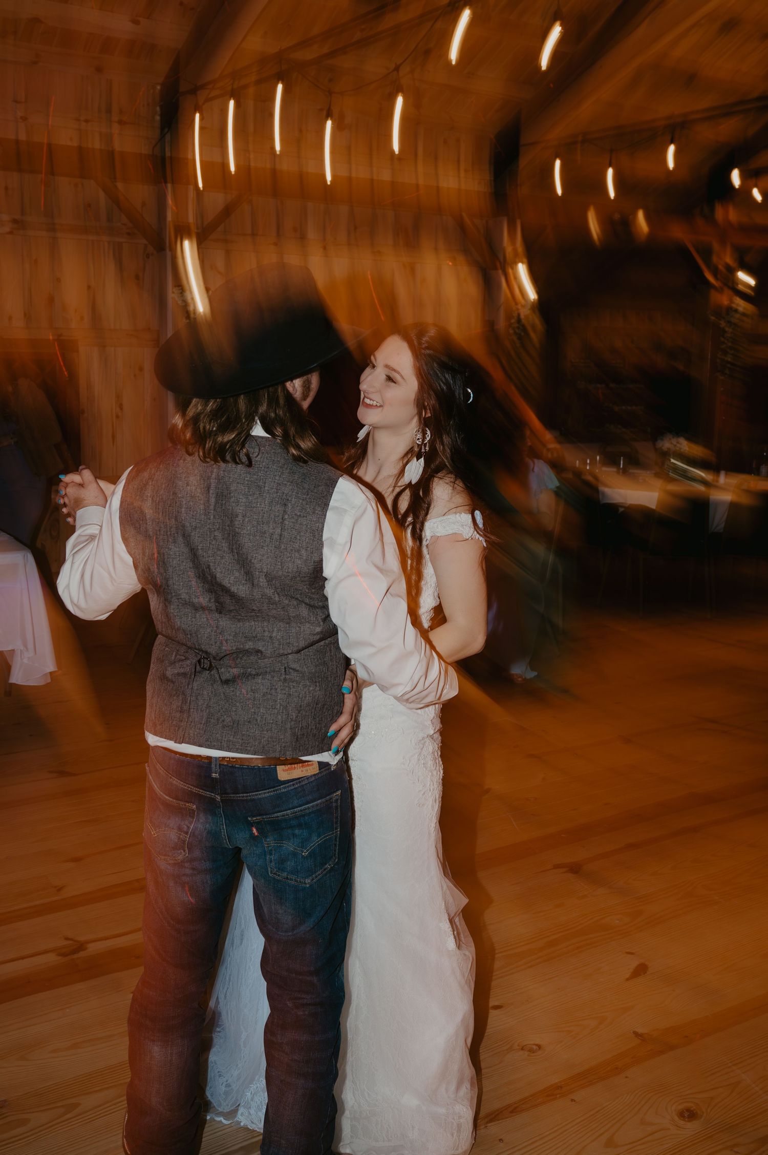 Bride and groom sharing first dance in warmly lit barn wedding venue with motion blur effect.