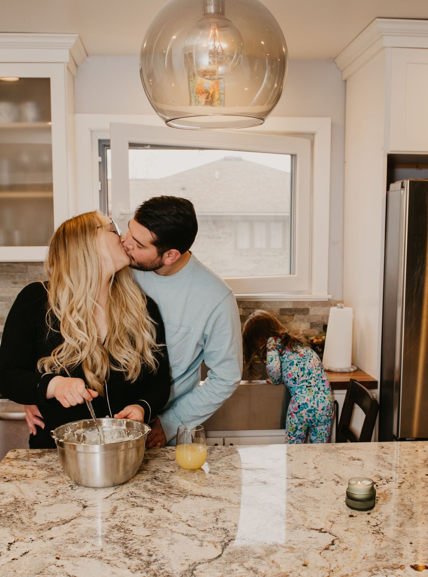 Couple sharing a kiss in modern kitchen while child plays in background.