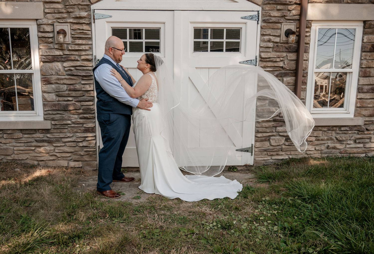 Bride and groom share a romantic moment outside a stone building, veil flowing in the breeze.