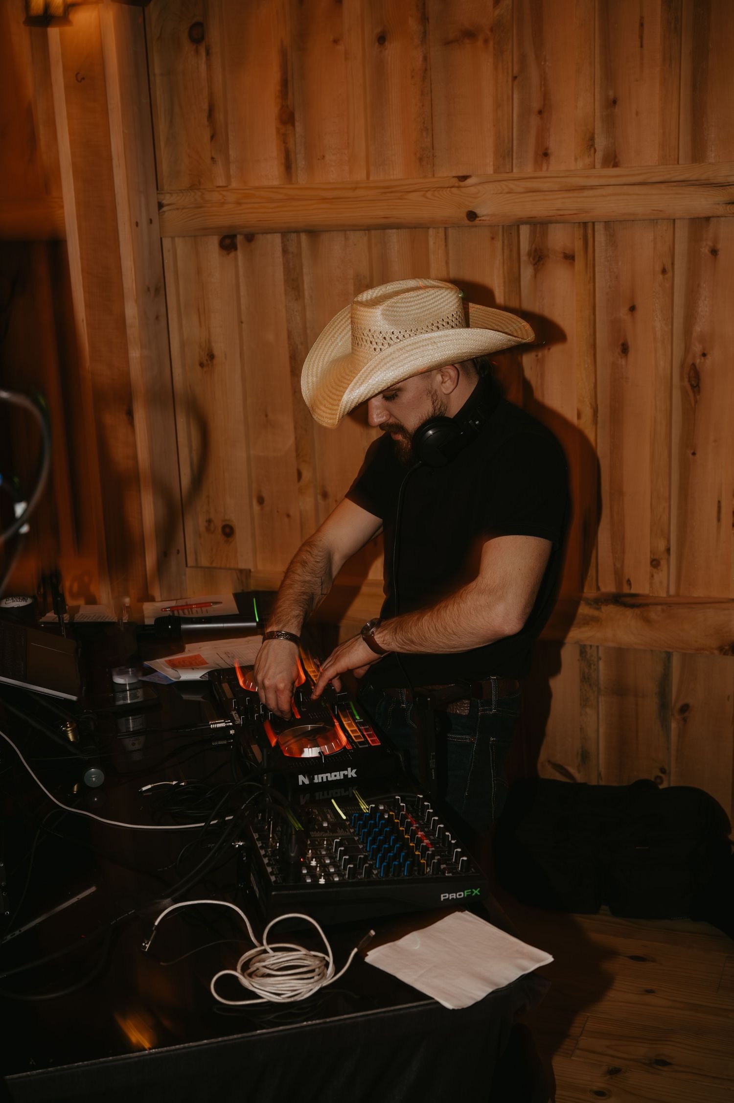 DJ in white cowboy hat performing at rustic barn wedding reception on wooden DJ equipment.