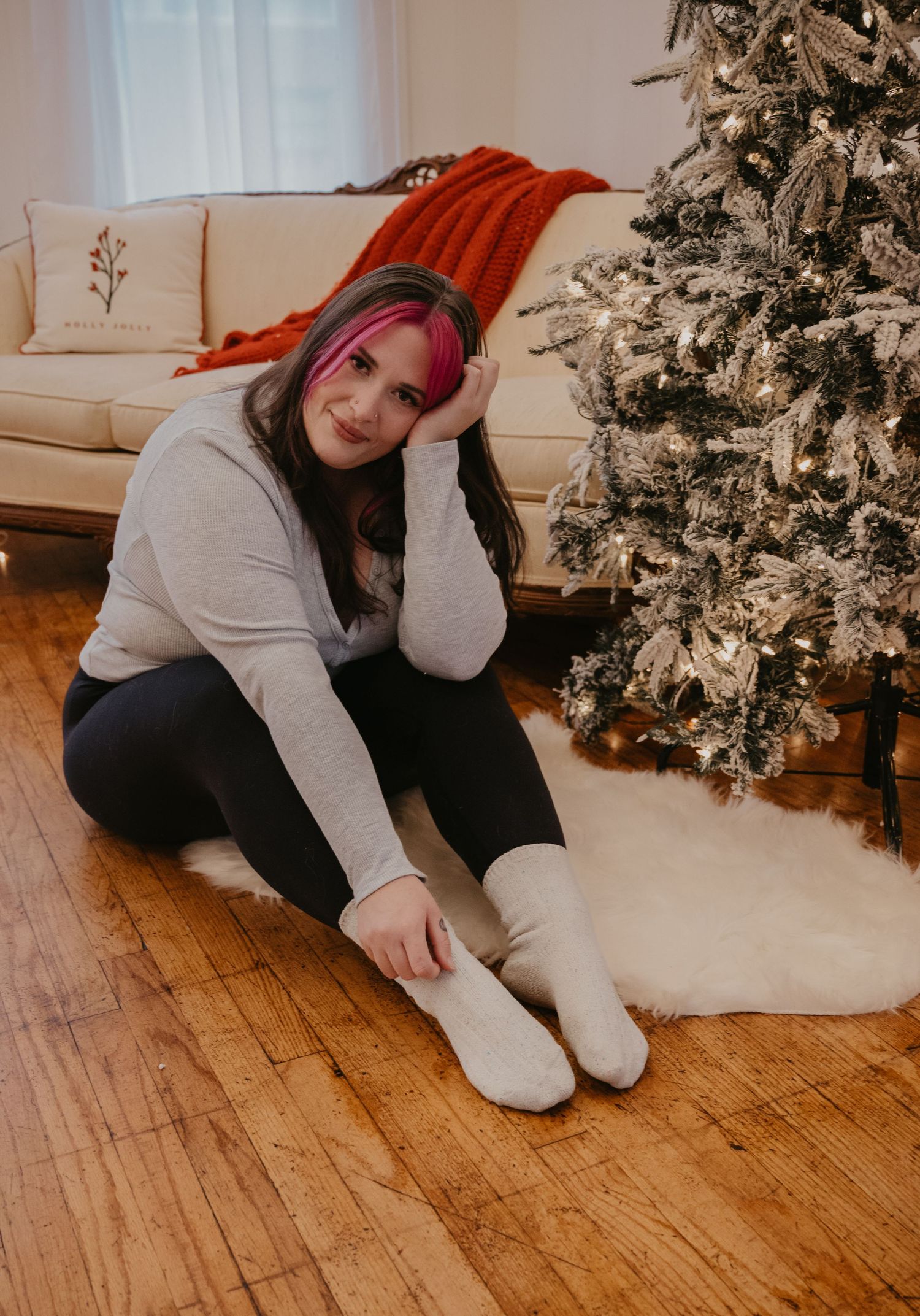 Young woman with pink highlighted hair sitting by flocked Christmas tree on hardwood floor.