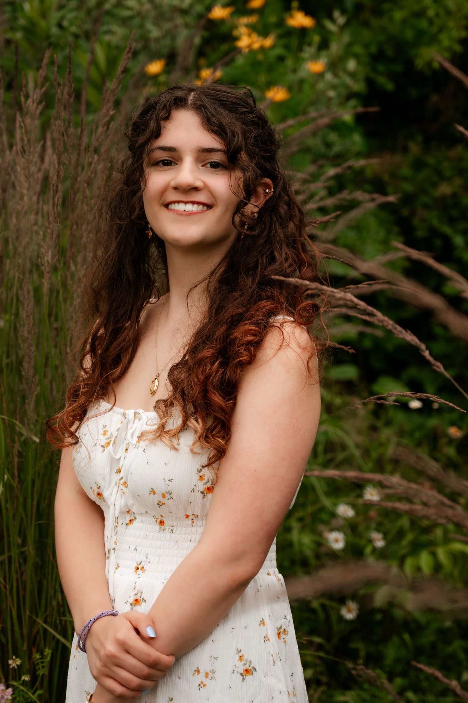 Smiling young woman with curly auburn hair in white floral dress poses among wildflowers and tall grasses.