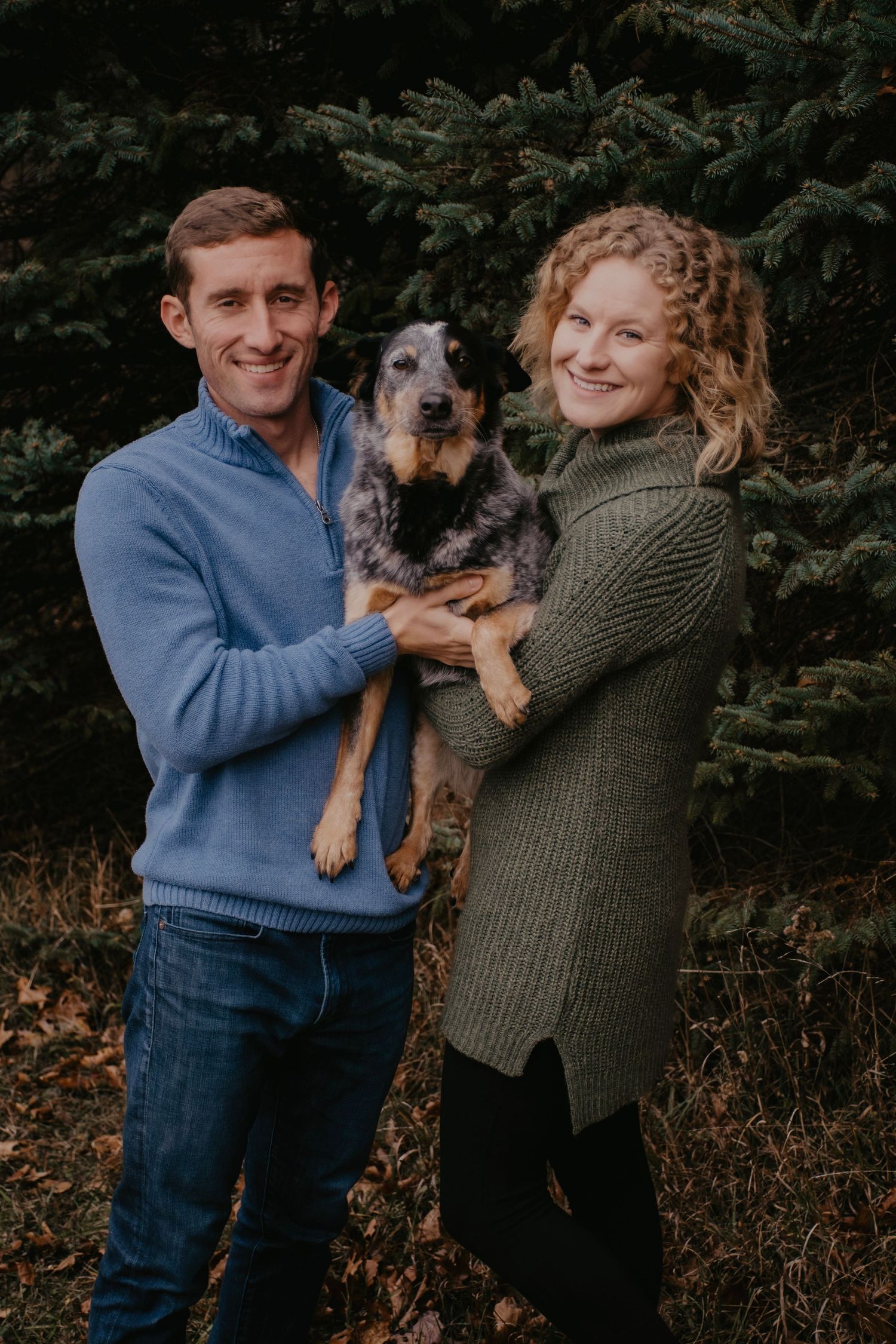 Smiling couple holding their blue heeler dog outdoors near evergreen trees.