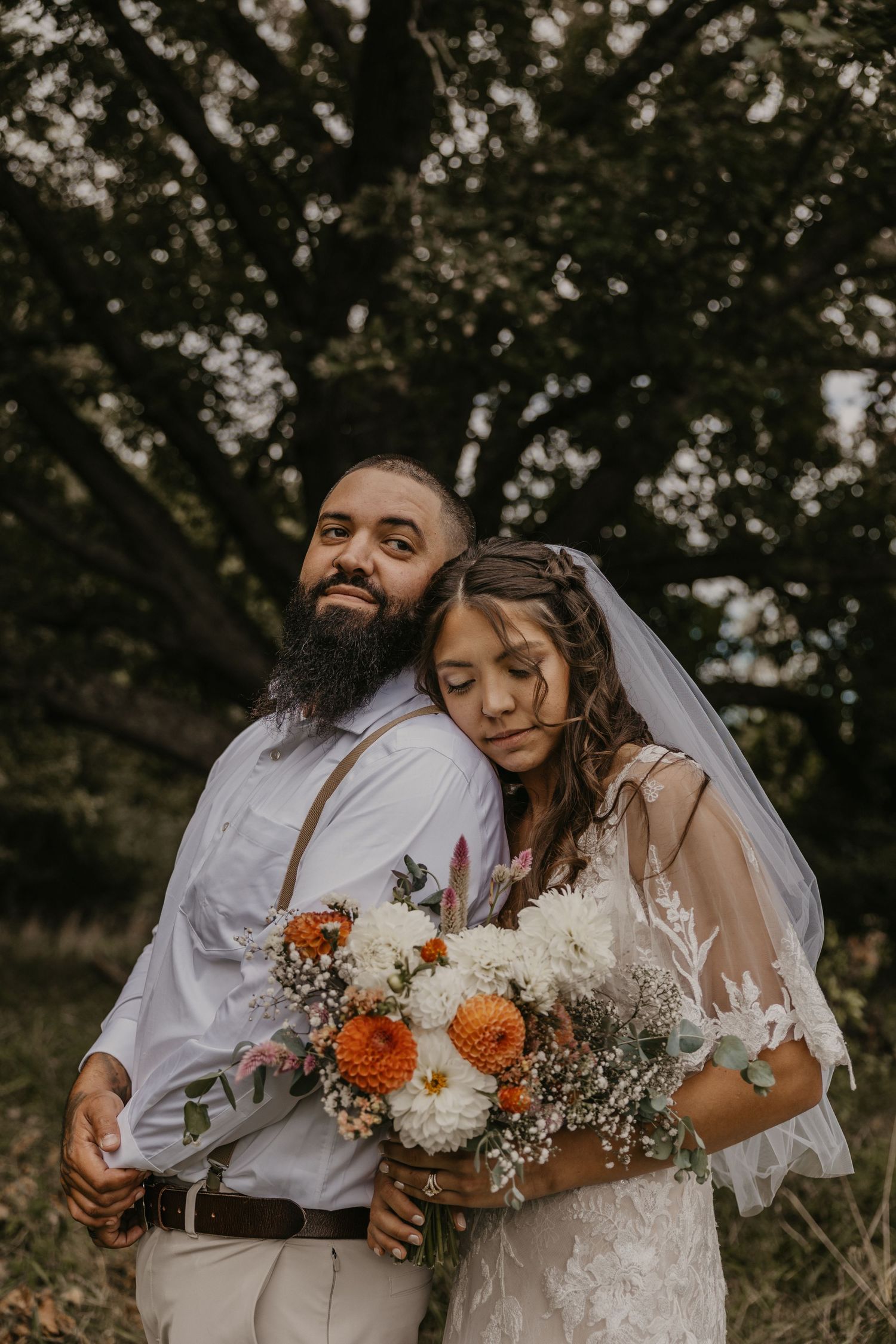 Bearded groom and bride in lace dress embrace back-to-back holding colorful wildflower bouquet outdoors.