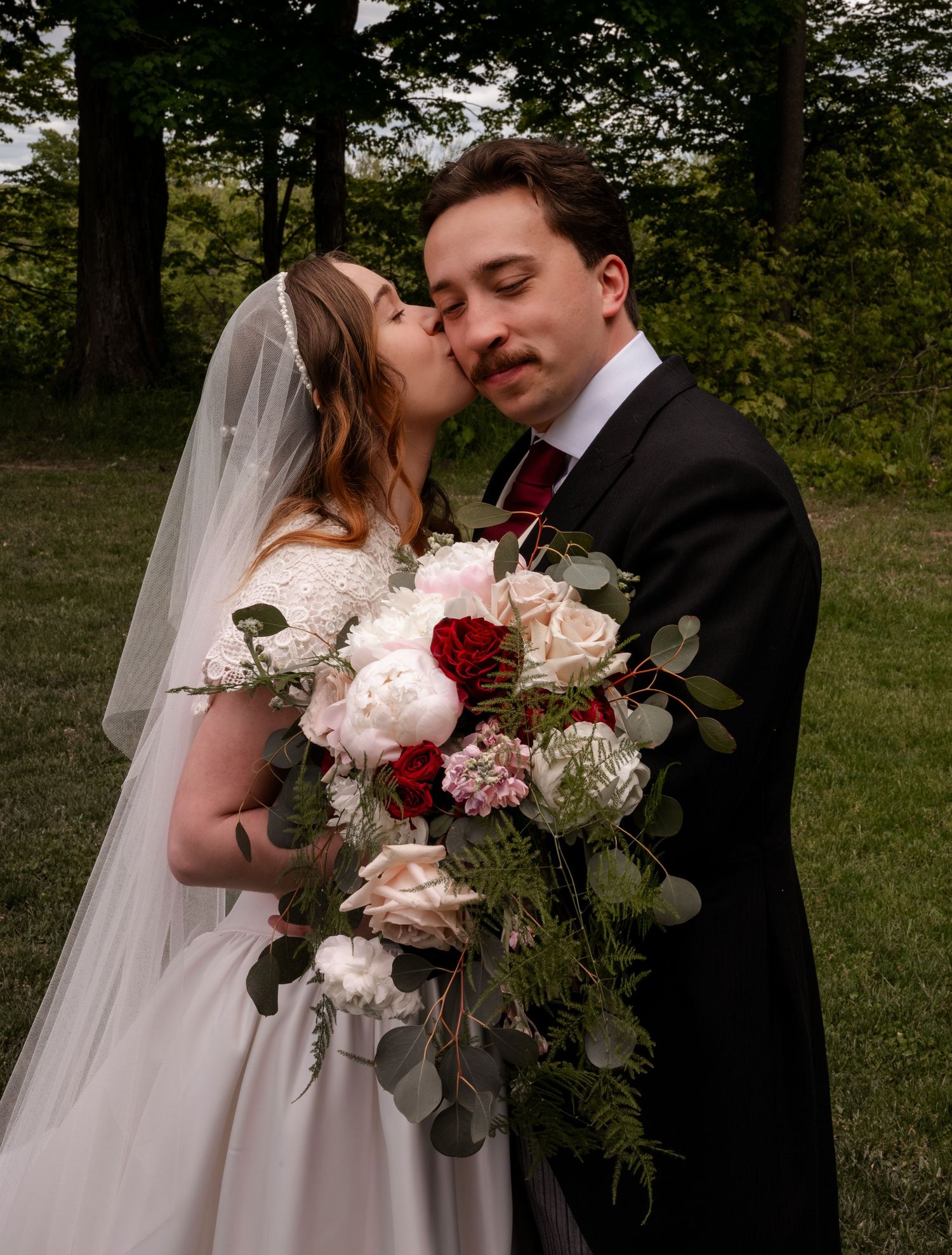 Bride kissing groom on cheek outdoors, holding lush bouquet of red and white flowers.