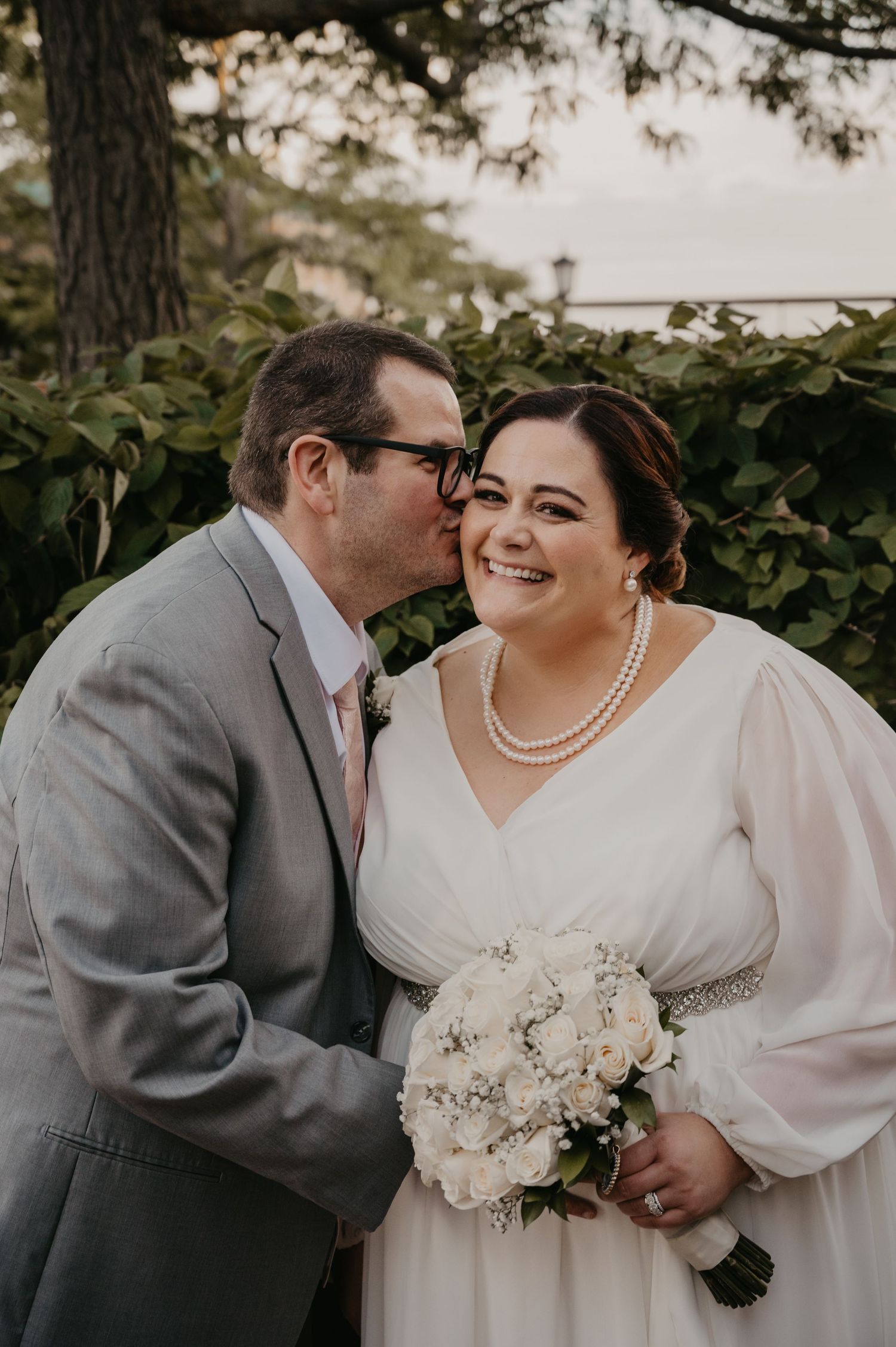 Groom in gray suit kisses bride on cheek as she smiles holding white bouquet outdoors.