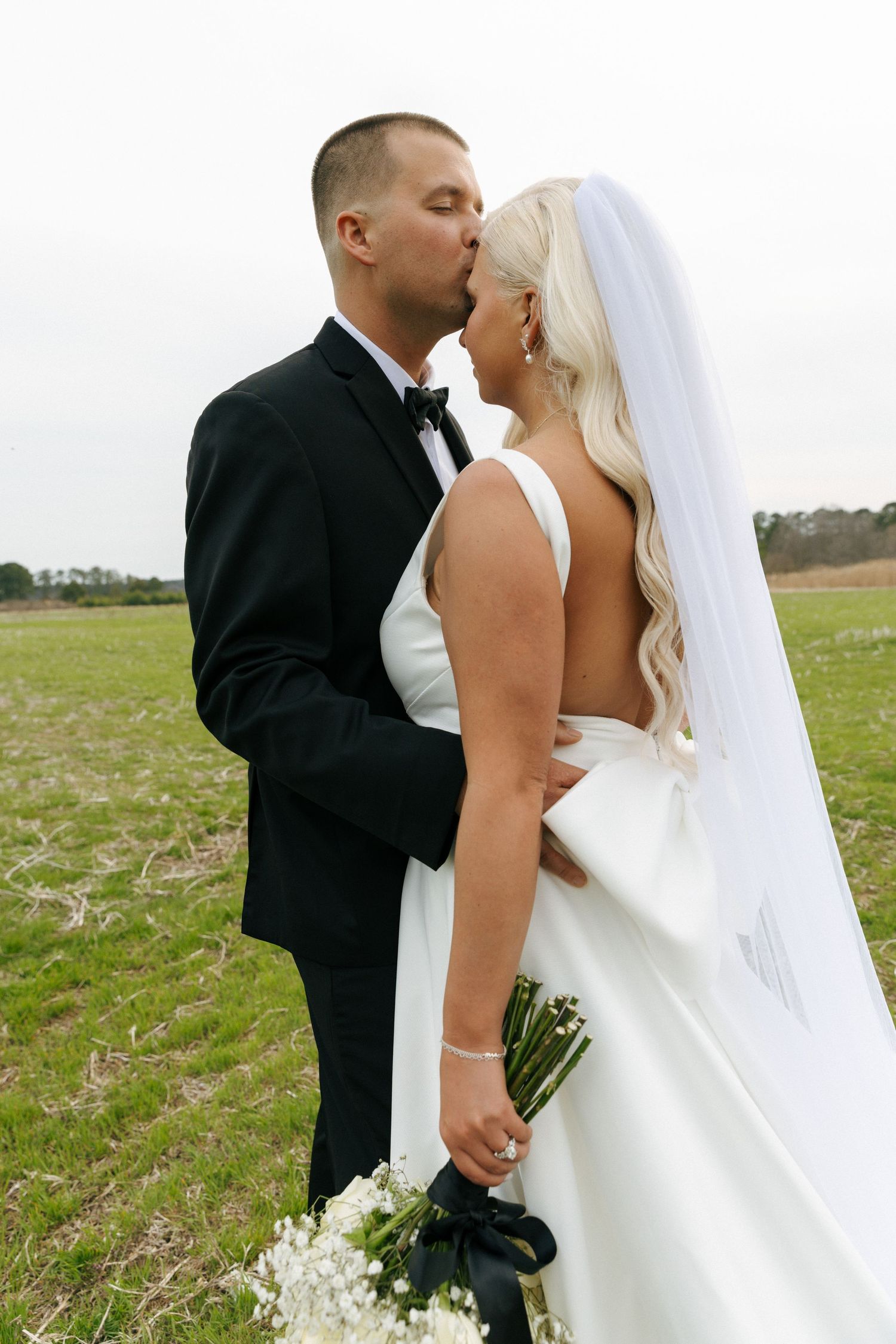 A tender moment captured between newlyweds in formal attire standing together in an open field with a bouquet.
