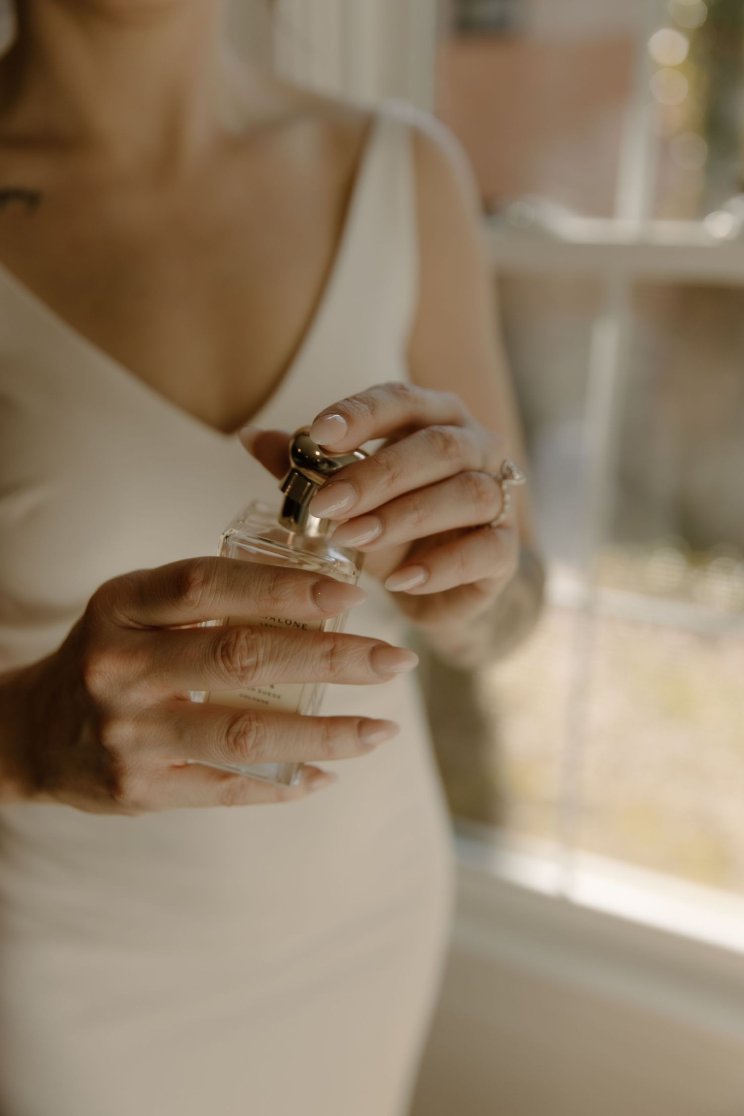 Hands gently holding a perfume bottle in soft natural light, wearing a white dress with delicate details.