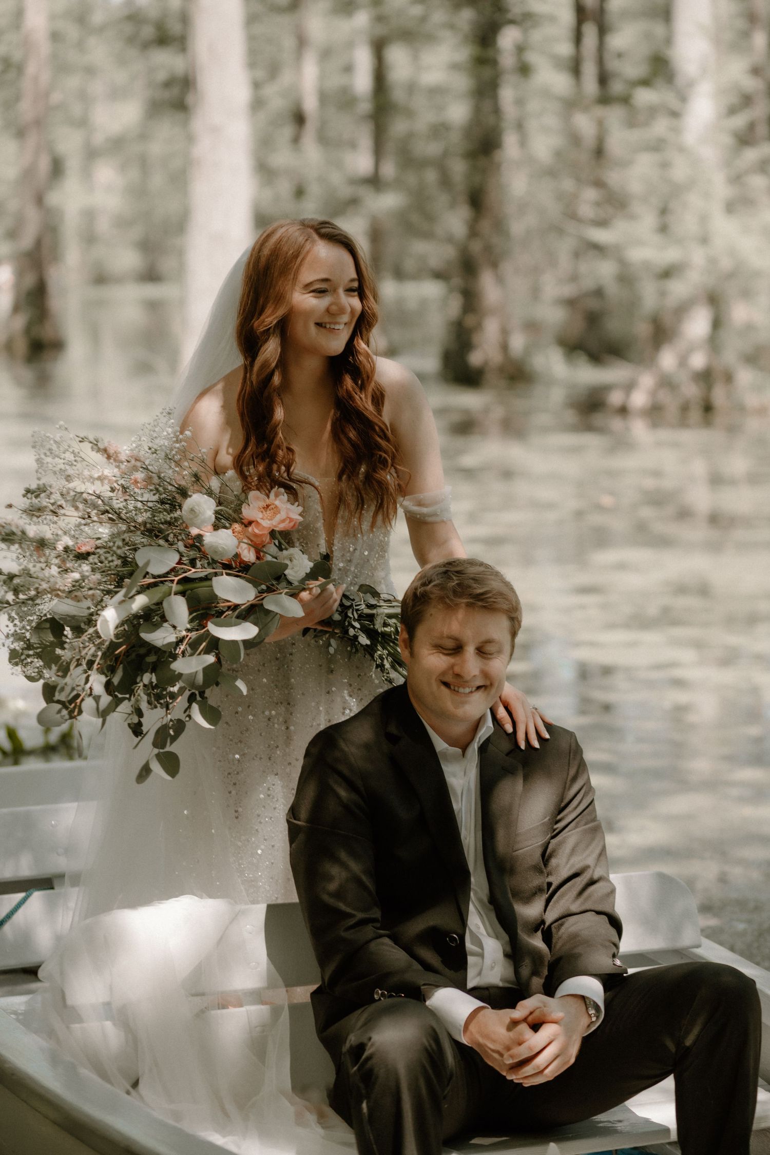 A romantic forest wedding photo with a couple seated on a rustic bench while holding a lush floral bouquet.