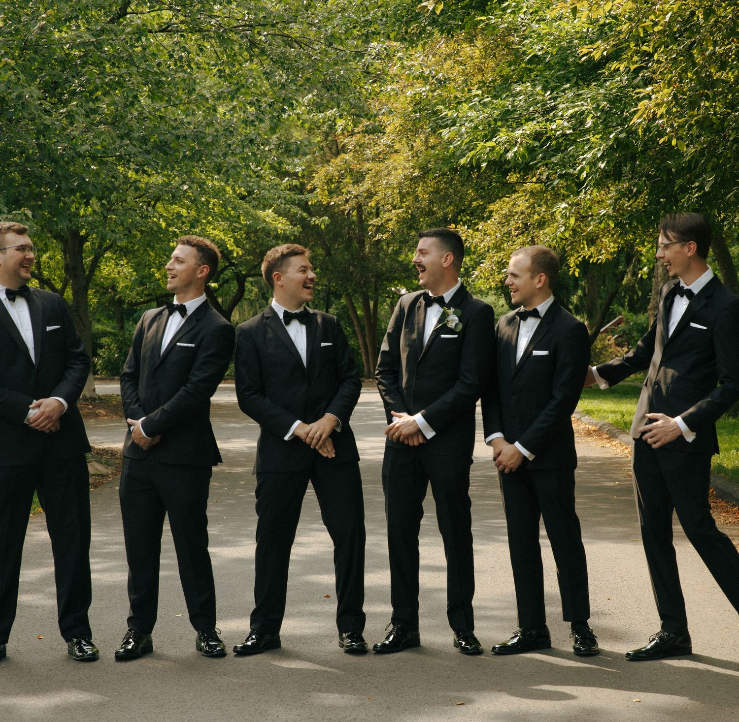 A row of groomsmen in matching black tuxedos and bowties pose casually outdoors under green trees.