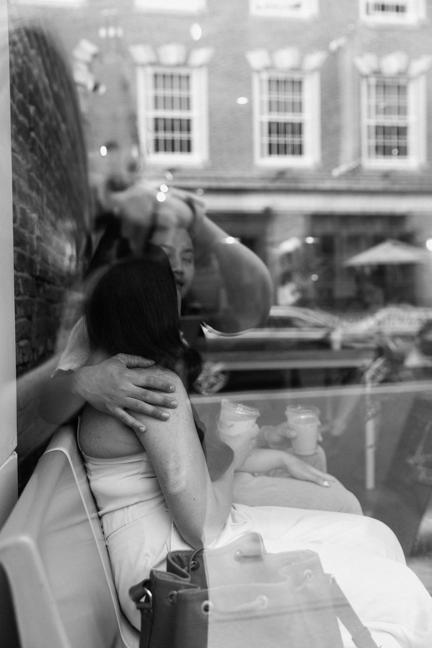 Artistic black and white photograph showing a person relaxing inside a cafe by a window with street reflections visible.