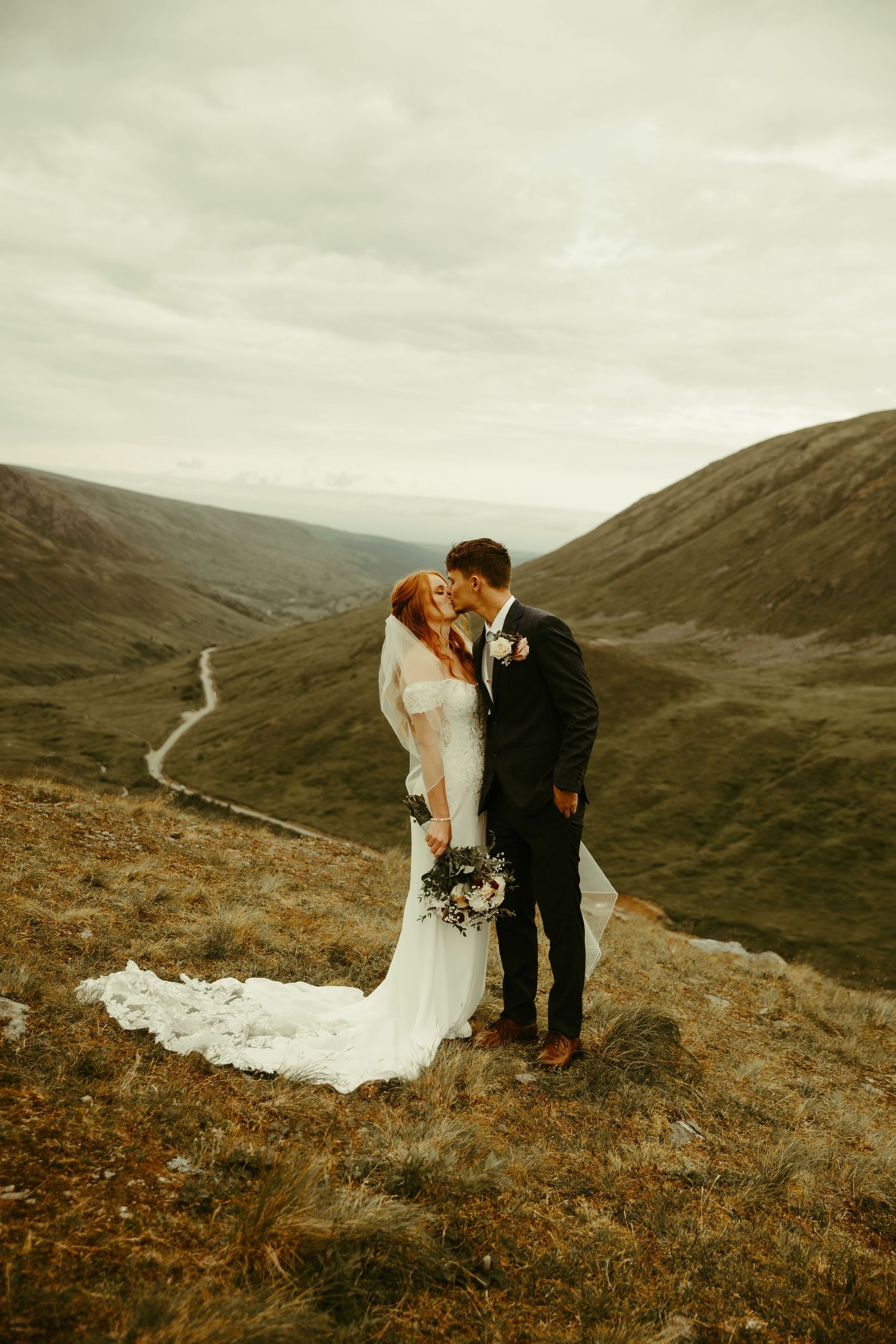 A wedding couple stands atop a scenic mountain valley with a winding road below on an overcast day in Scotland.