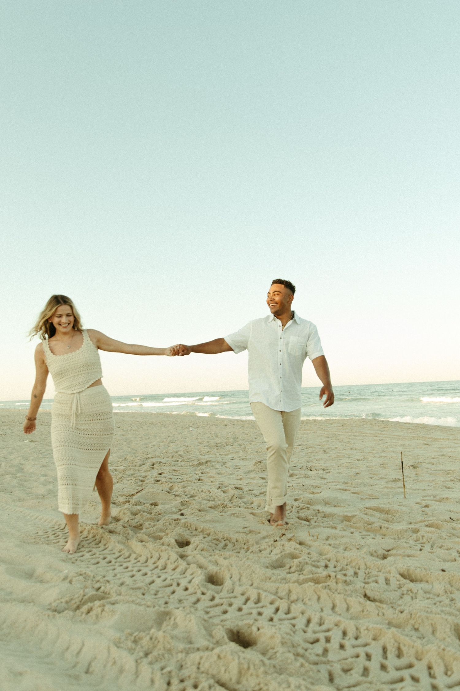 A couple in white outfits walks hand in hand along a sandy beach at sunset with ocean waves in the background.