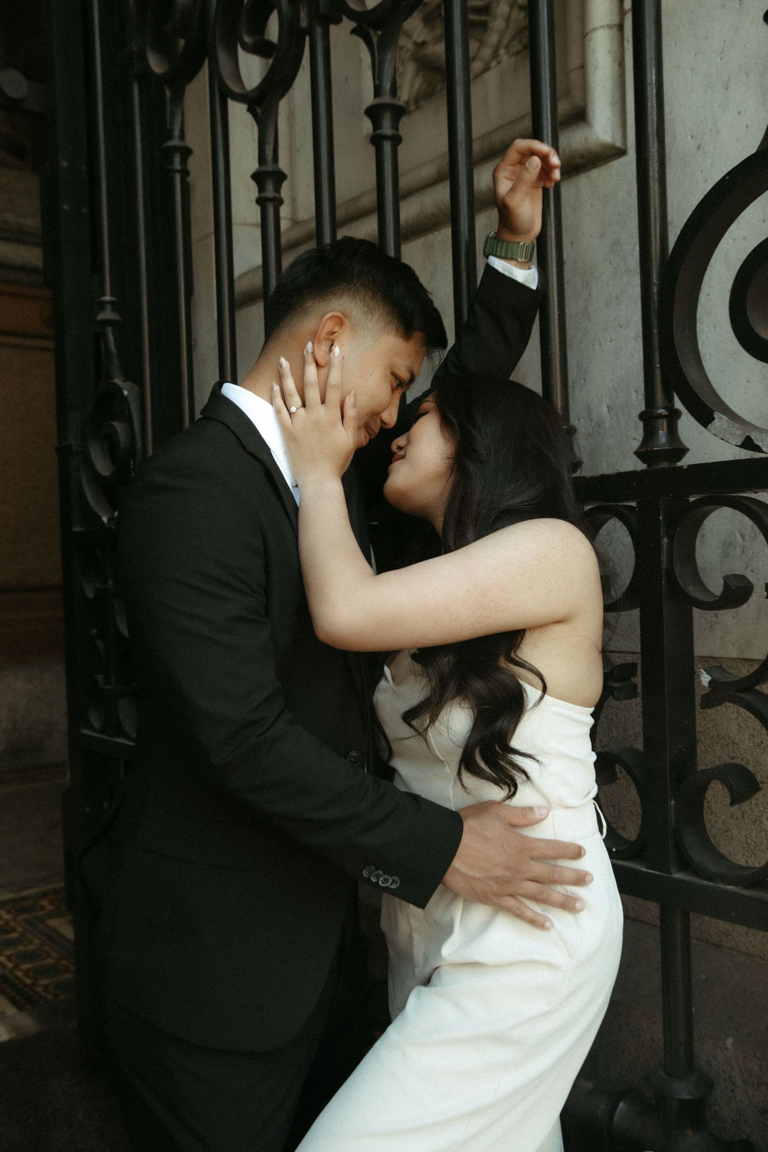 A romantic couple in formal wedding attire embrace against an ornate iron gate in a dimly lit setting.