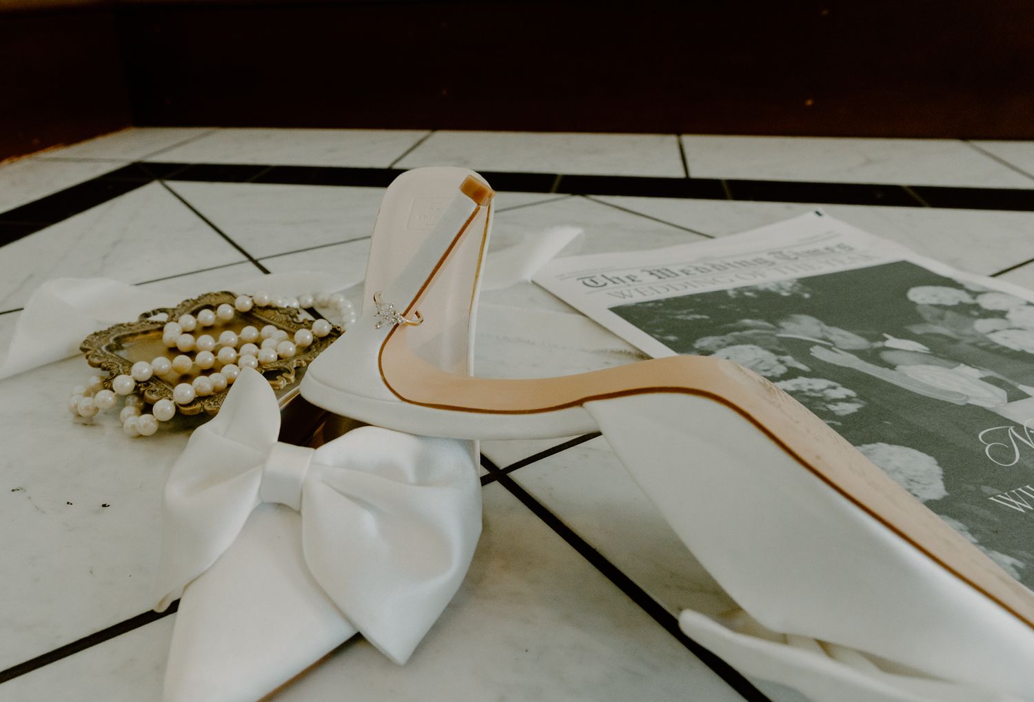 White high heel shoe, ribbon bow, and golden jewelry arranged on a geometric tiled surface in soft vintage tones.