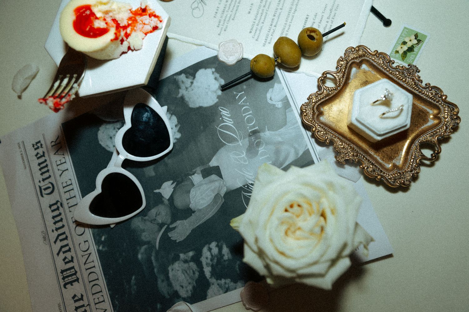 Wedding themed flat lay with heart sunglasses, white rose, and decorative frame on a vintage newspaper background.