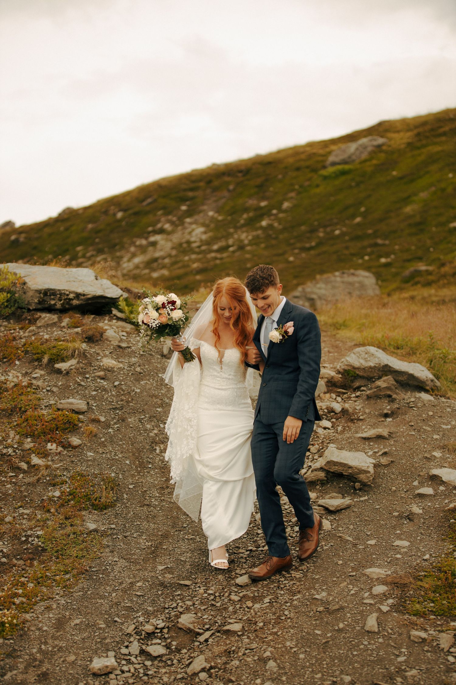 A couple in wedding attire walking together along a rocky mountain path against a scenic highland backdrop.