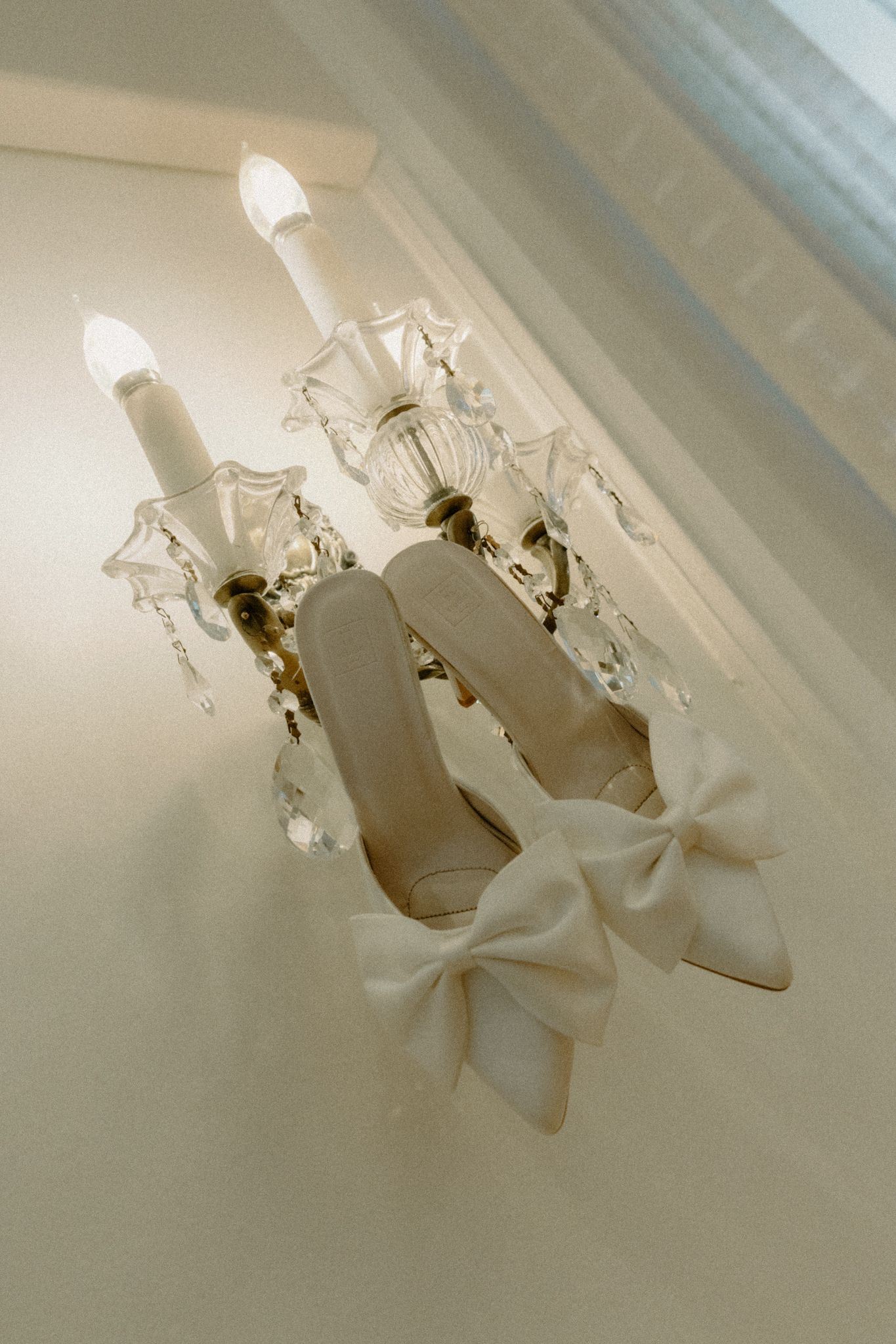 White bridal shoes with bows sit elegantly beneath a crystal chandelier in soft lighting.