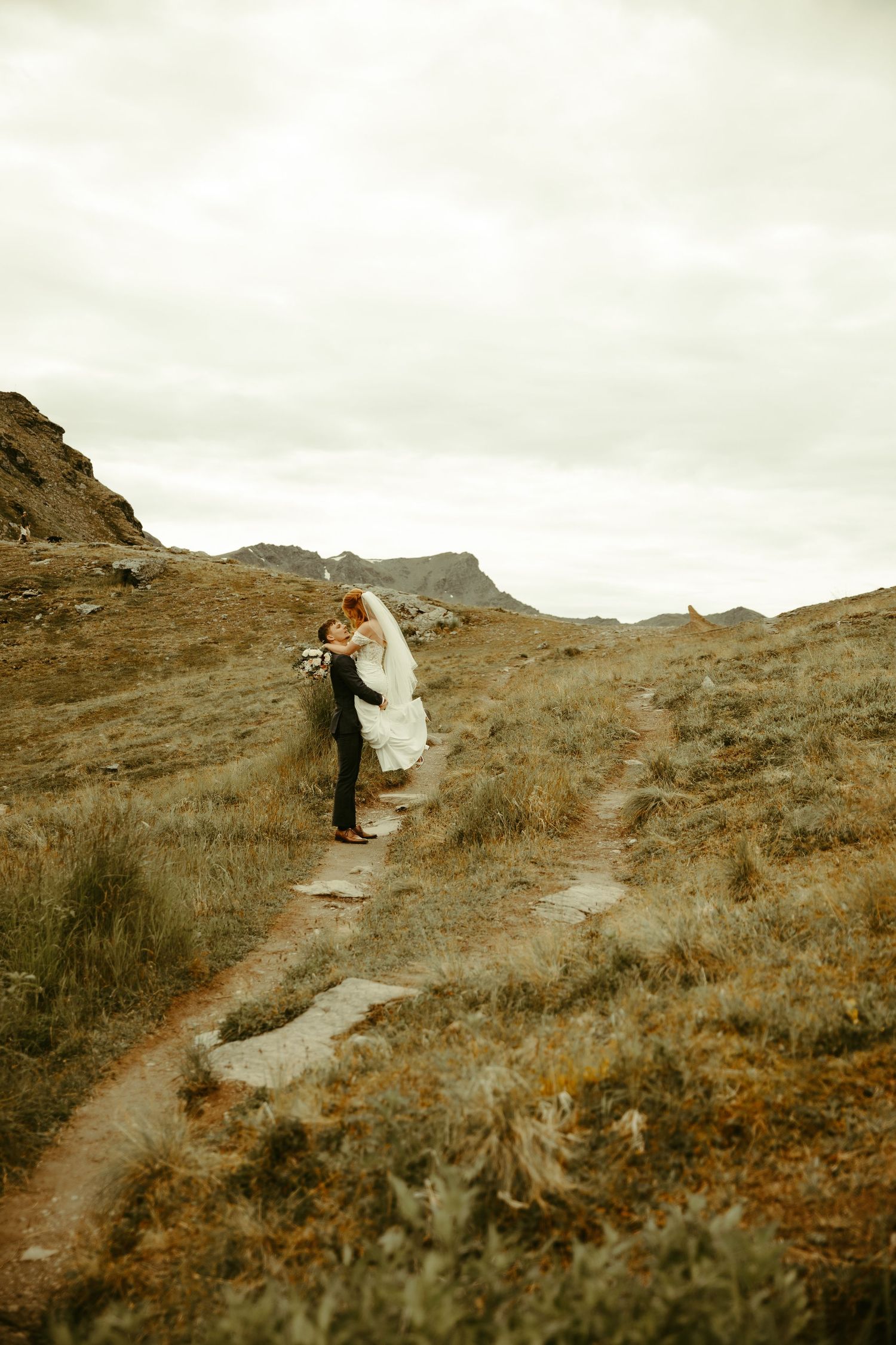 A couple embraces on a winding mountain trail surrounded by rugged terrain under an overcast sky.