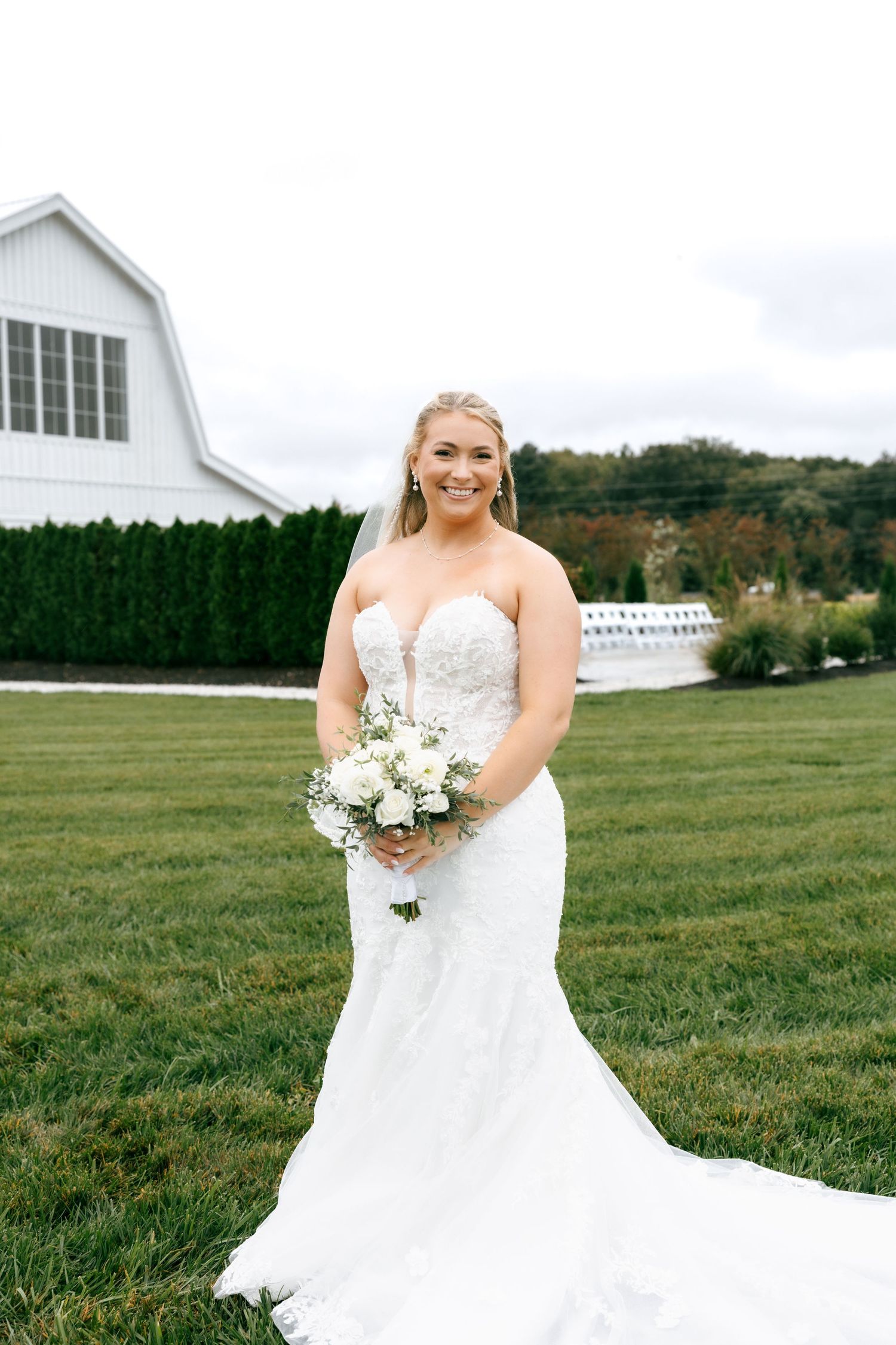 A bride in a flowing white wedding dress poses in front of a white barn on a green lawn with rolling hills.