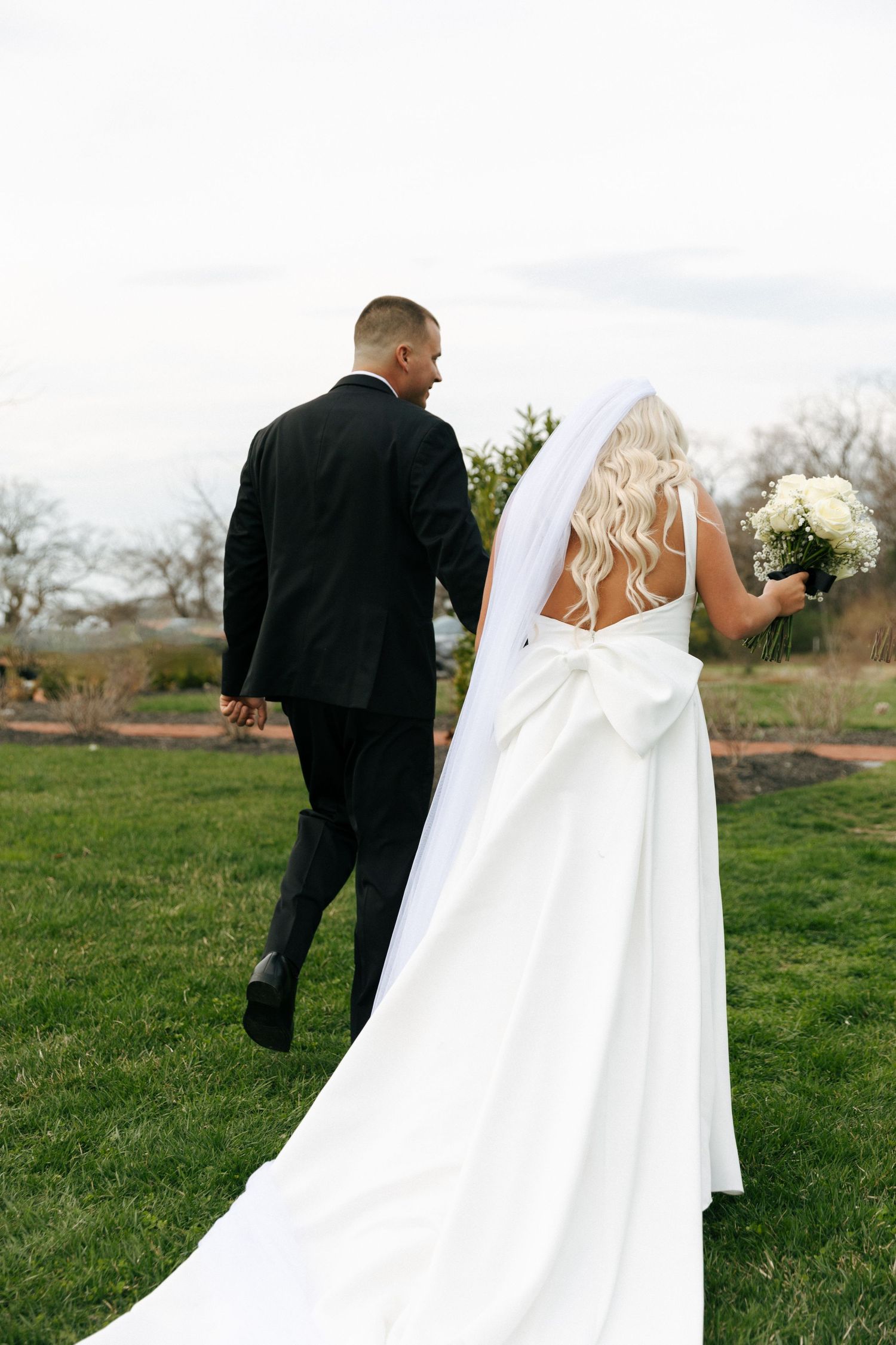A couple in wedding attire walking on grass, with the bride wearing a long flowing white dress and holding a bouquet.