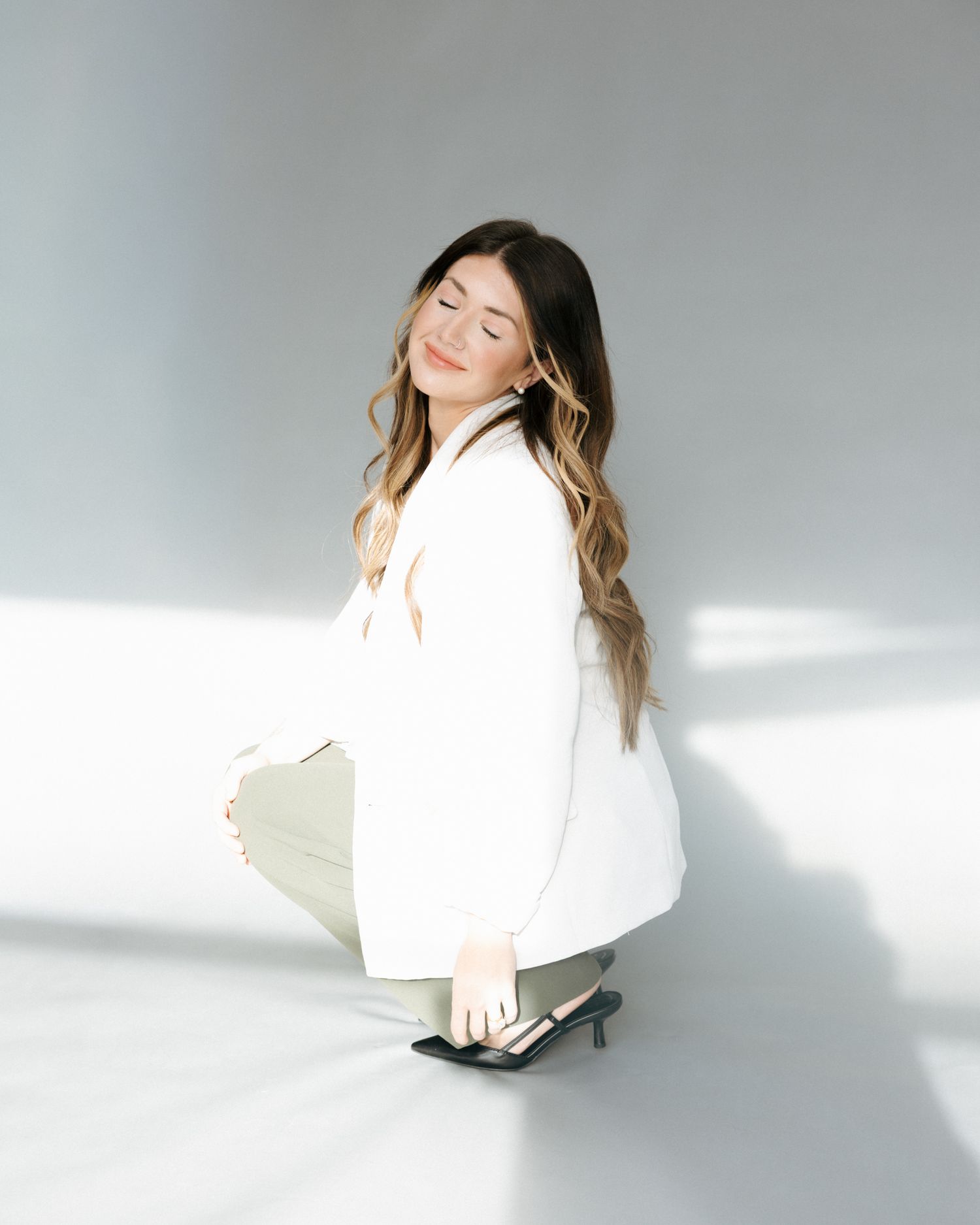 Person in a white sweater crouches against a light gray background with dramatic shadows and lighting.